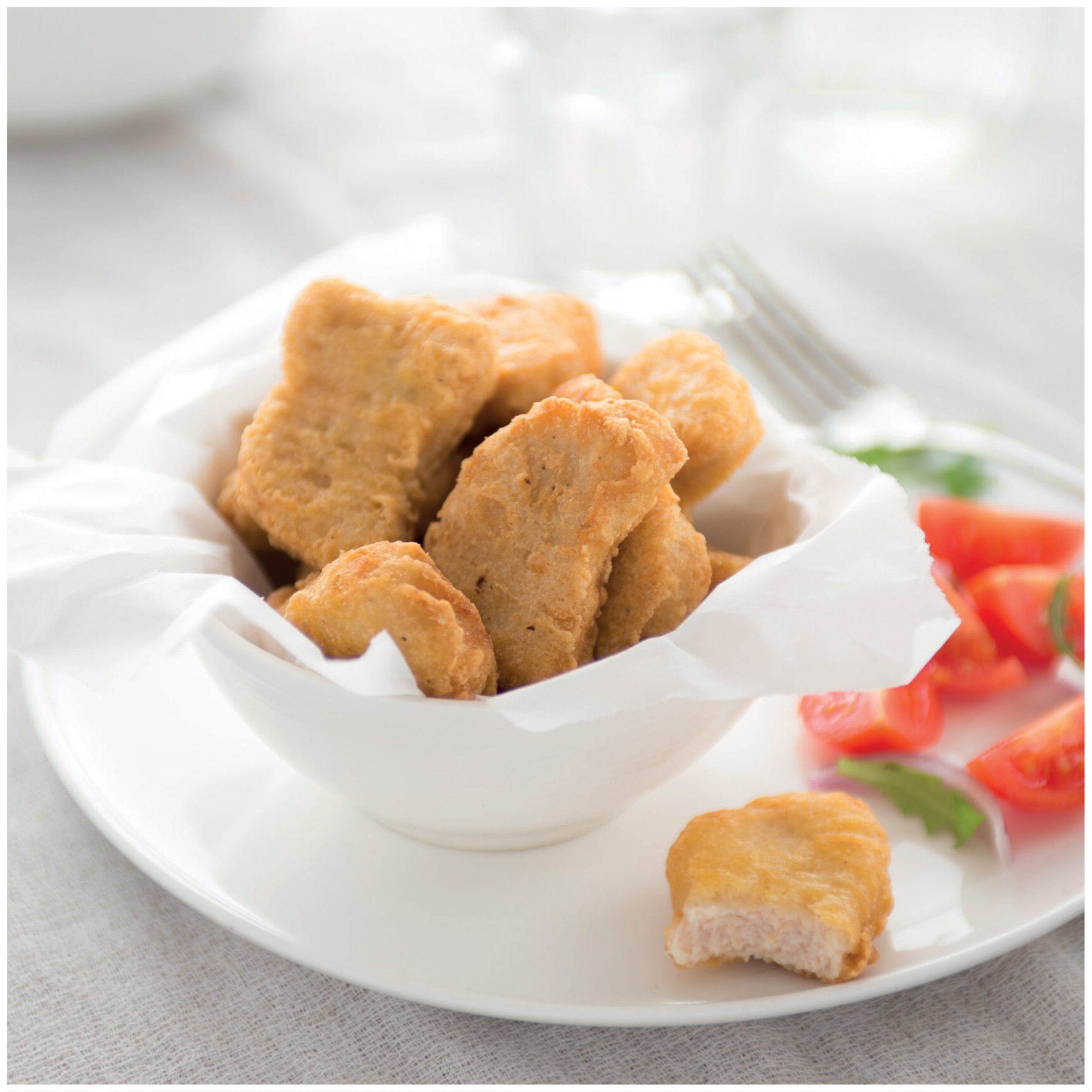 Nuggets di pollo dorati in una ciotola bianca con pomodori e lattuga su un piatto.