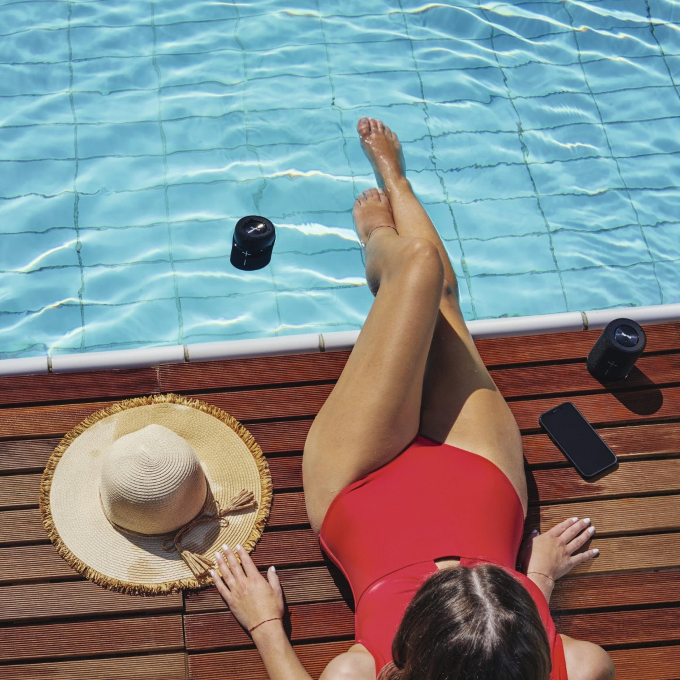 Kobieta w czerwonym stroju kąpielowym przy brzegu basenu, z kapeluszem, telefonem i głośnikami.