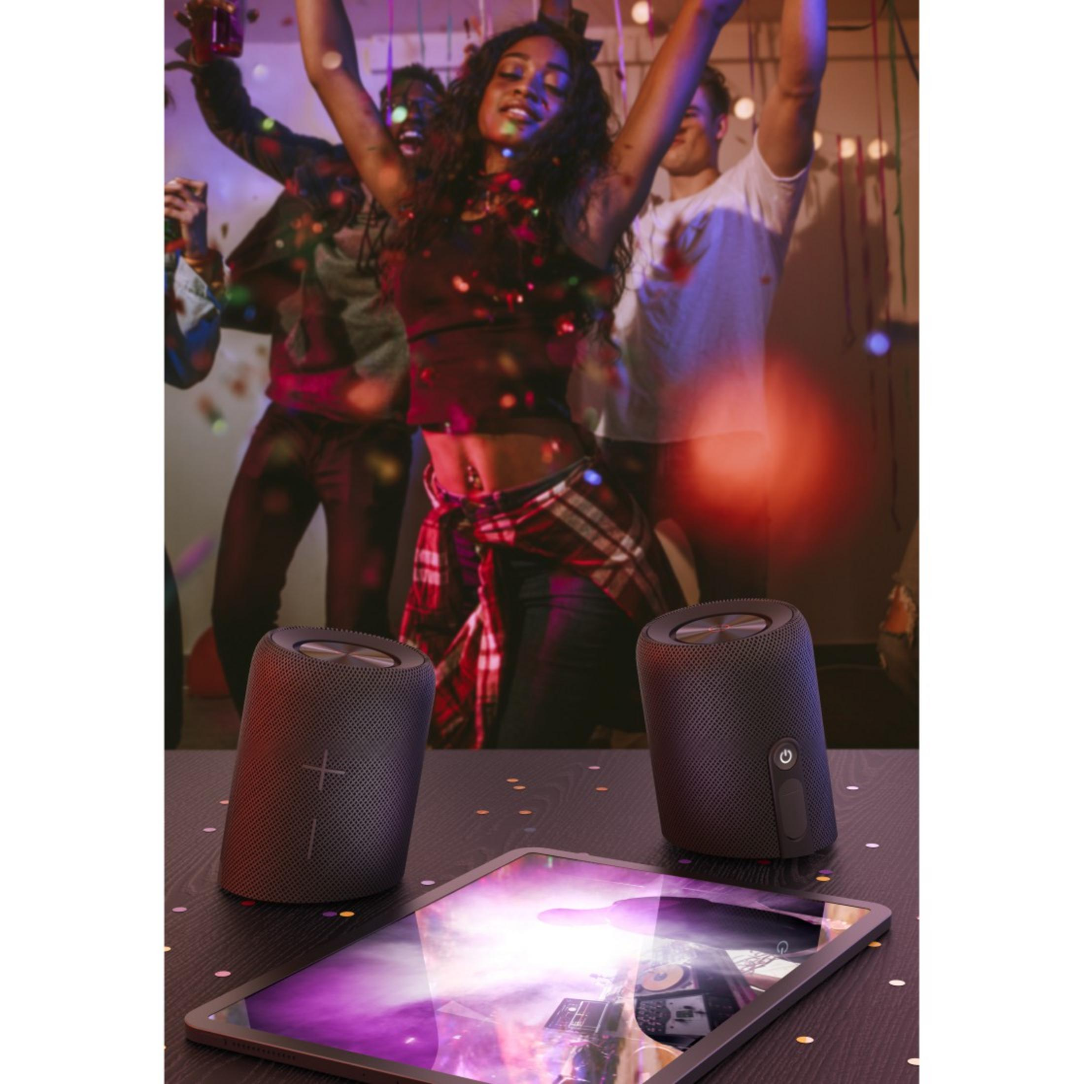 Zwei schwarze Lautsprecher und ein Tablet stehen auf einem Holztisch. Menschen tanzen im Hintergrund bei einer Party.