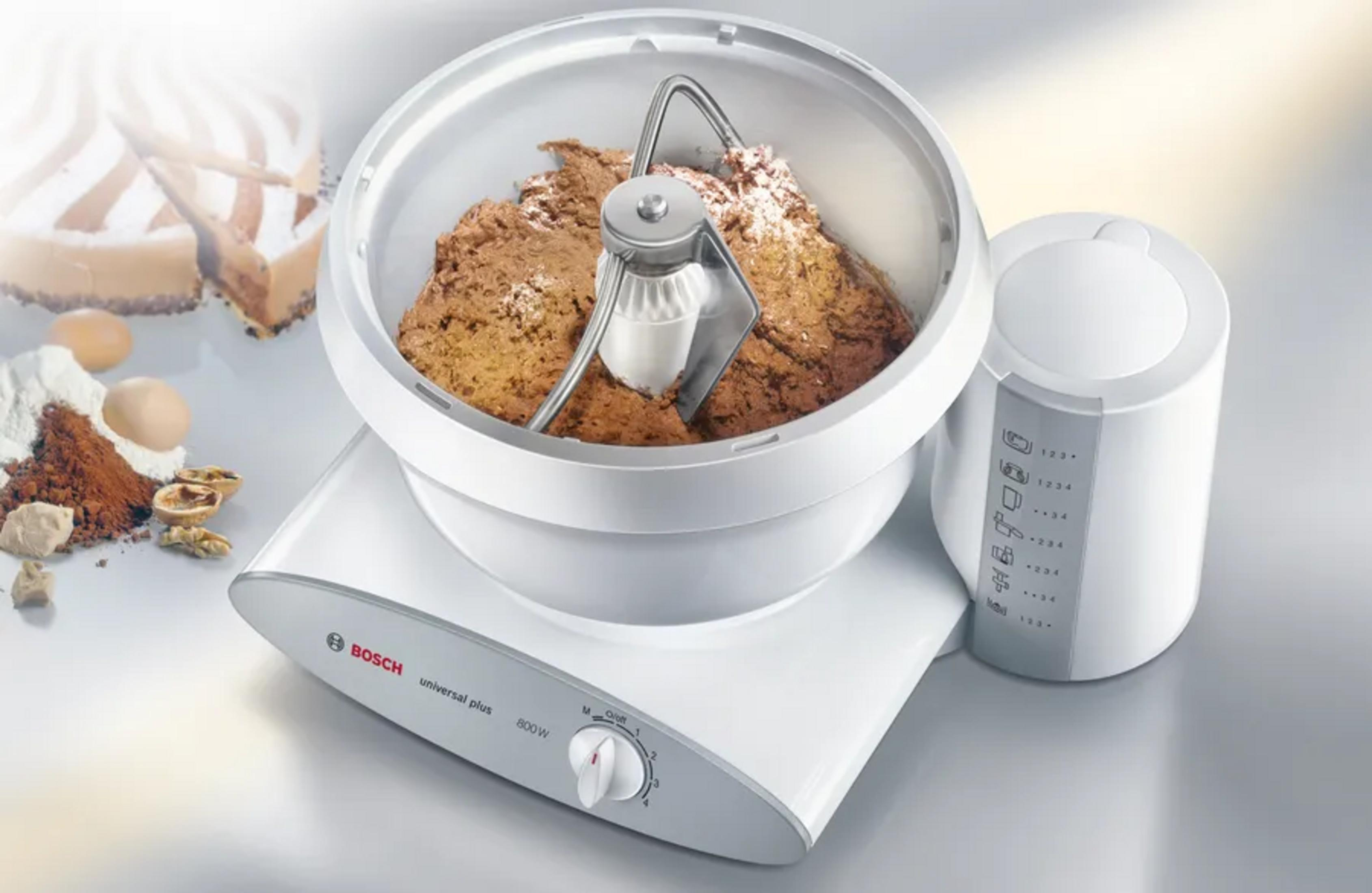 Een witte keukenmixer met beslag in de kom en taart op de achtergrond.