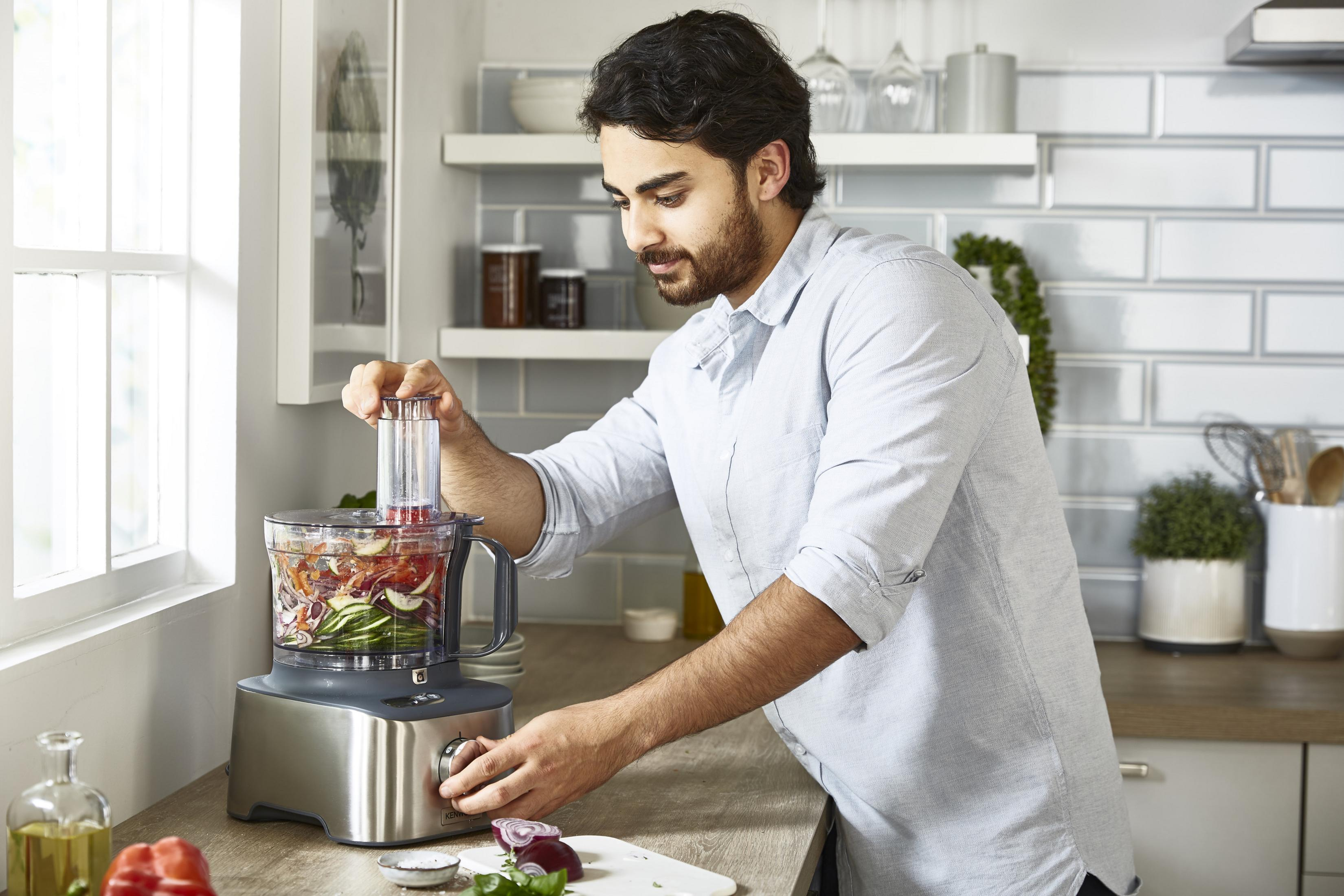 Mężczyzna w jasnoniebieskiej koszuli używa robota kuchennego z warzywami.