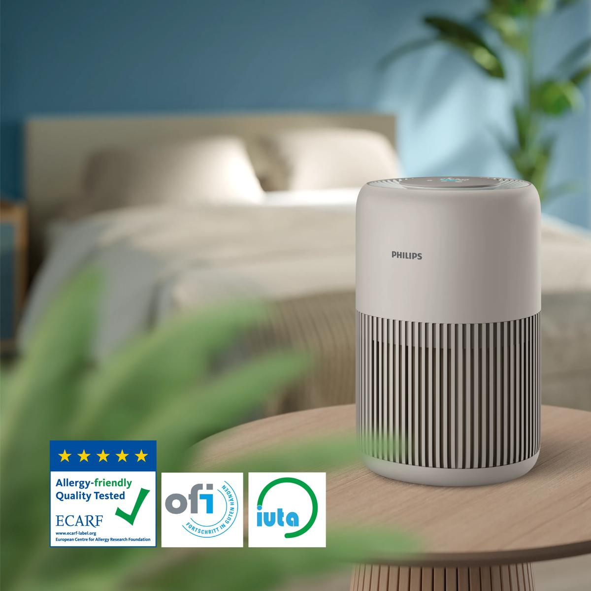 A white Philips air purifier sits on a wooden table in front of a bed.