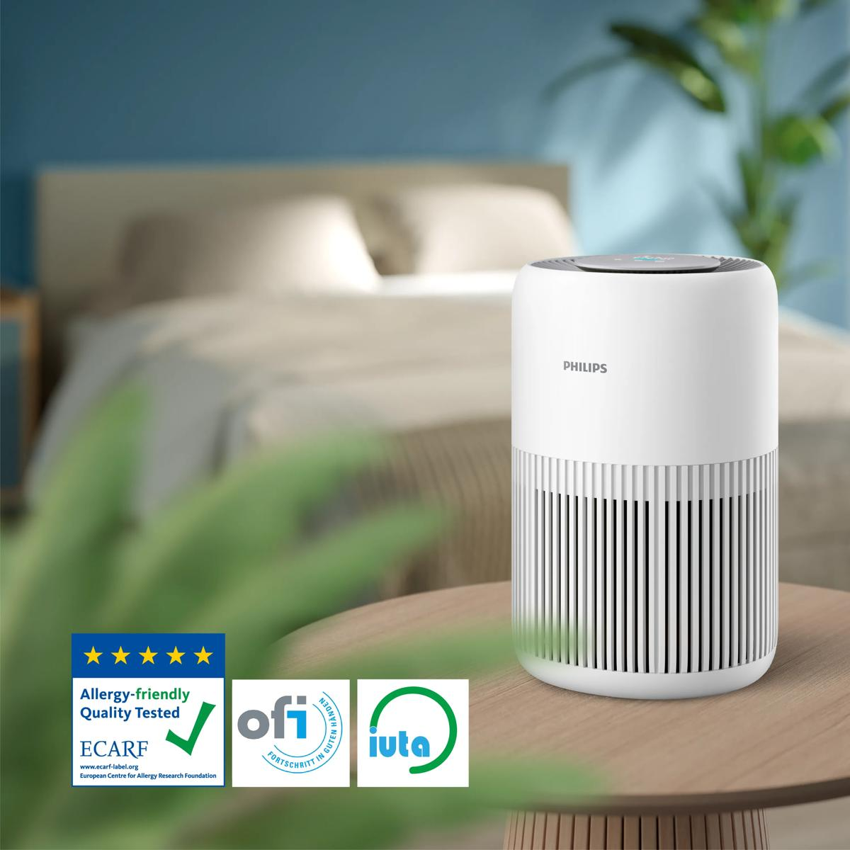 Un purificador de aire blanco Philips está sobre una mesa de madera frente a una cama desenfocada.