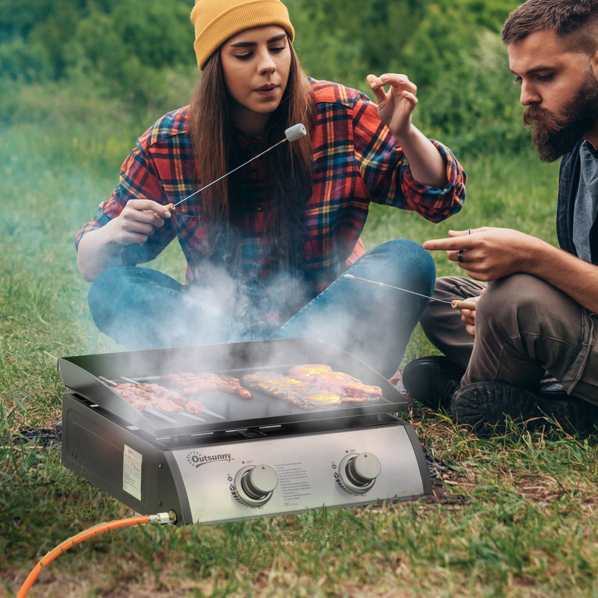 Un uomo e una donna seduti sull'erba, grigliano carne su un barbecue portatile.