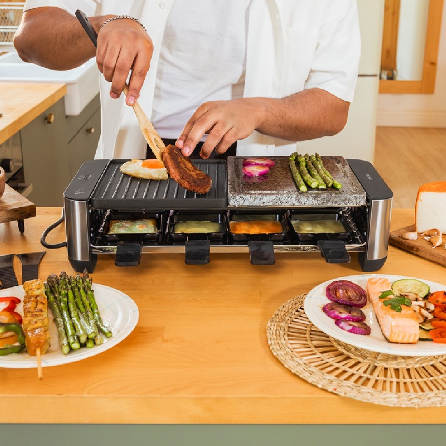 Ein Mann kocht auf einem Raclette-Grill mit Spargel, Ei und Fleisch, dazu Beilagen.