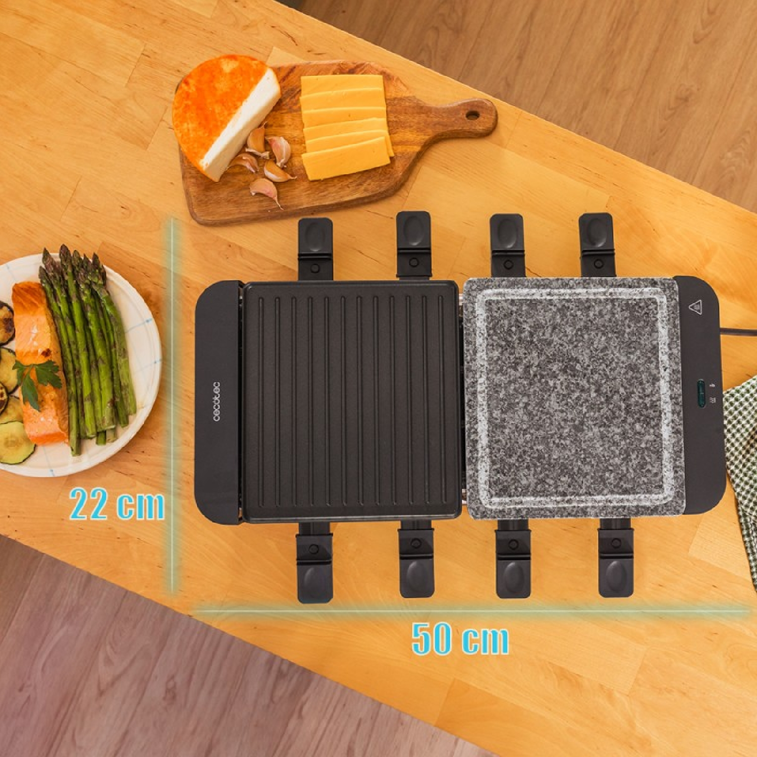 Ein Raclettegerät mit Stein- und Grillplatte steht auf einem Holztisch mit Lebensmitteln.