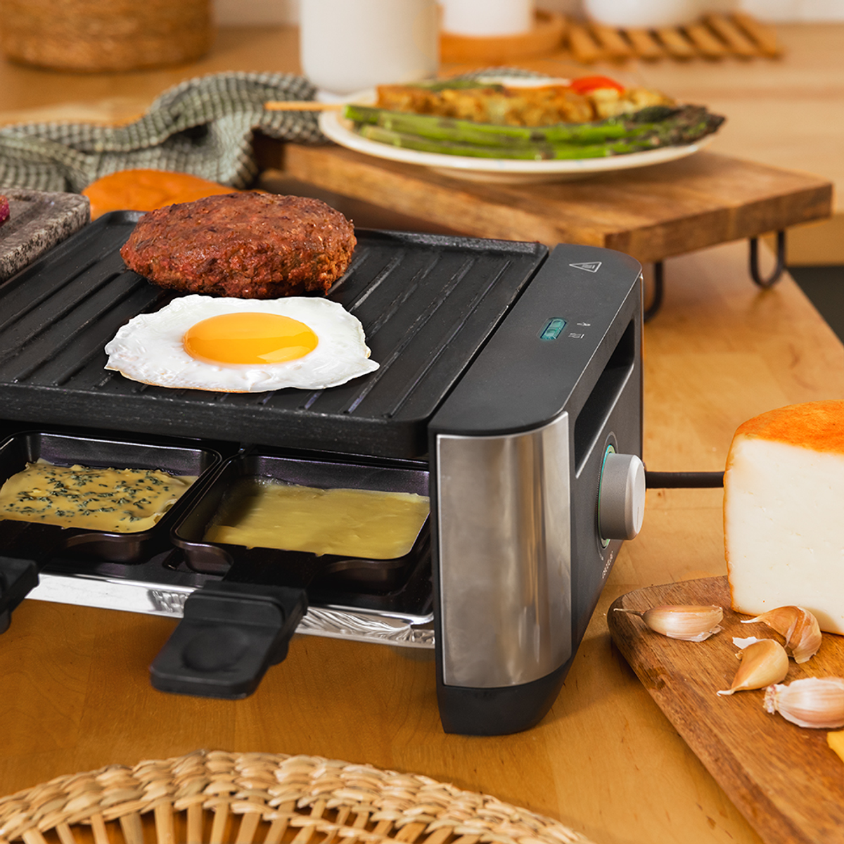 Una griglia per raclette cuoce un hamburger e un uovo fritto, con padelle per formaggio sotto.