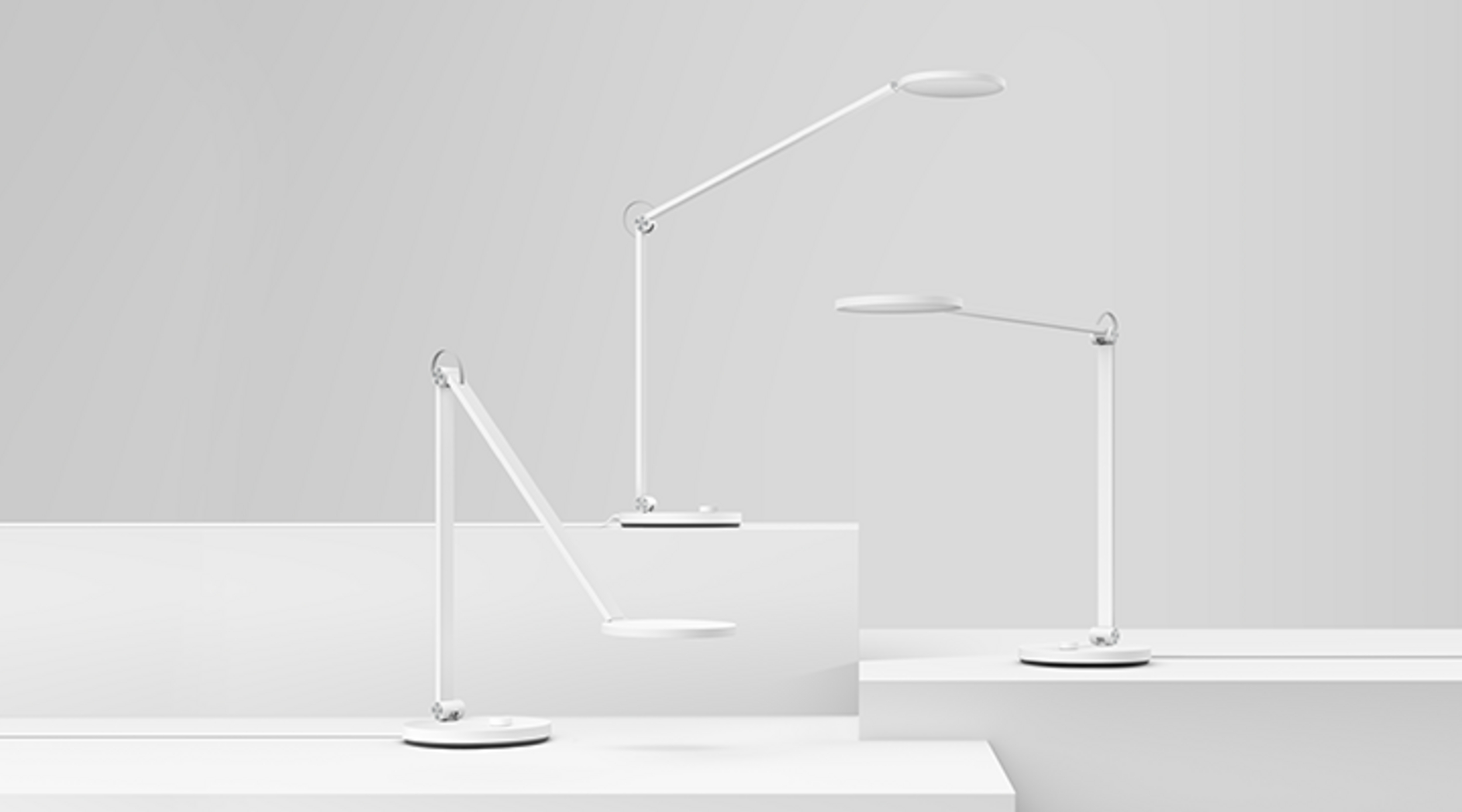 Trzy białe lampki biurkowe na białych platformach na szarym tle.