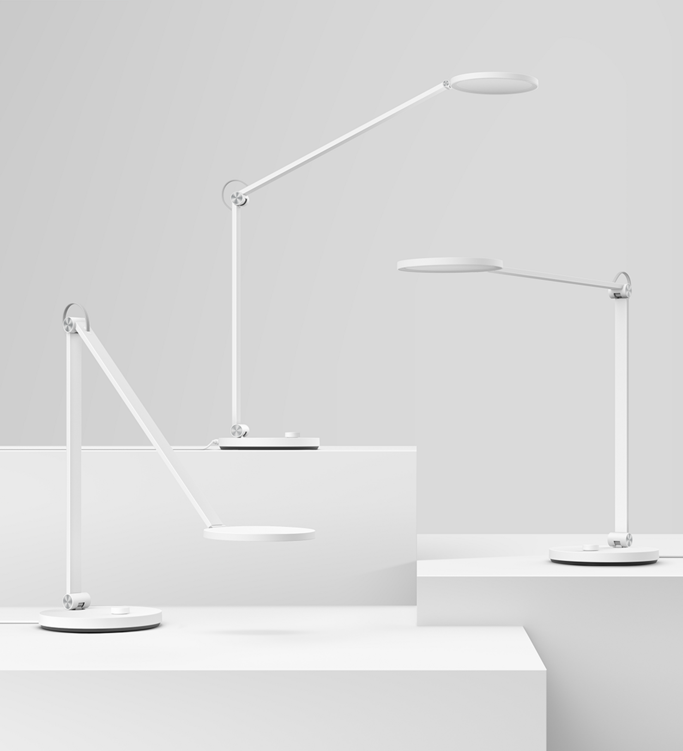 Trzy białe, nowoczesne lampki biurkowe na białych postumentach na szarym tle.