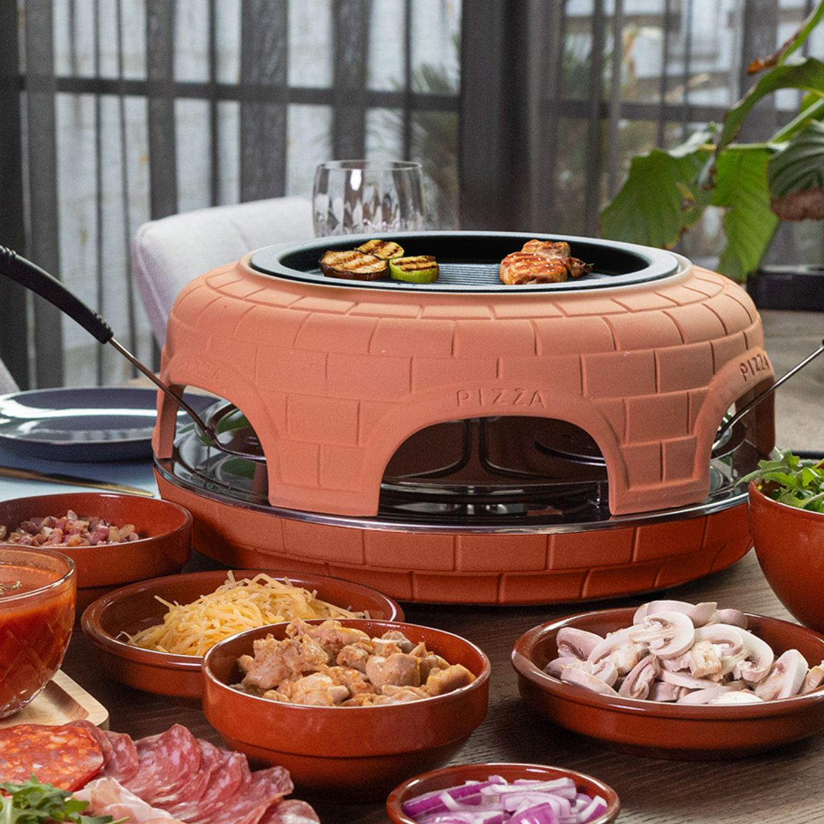 Een terracotta pizzaoven met gegrild vlees en courgette, omringd door schalen met ingrediënten.