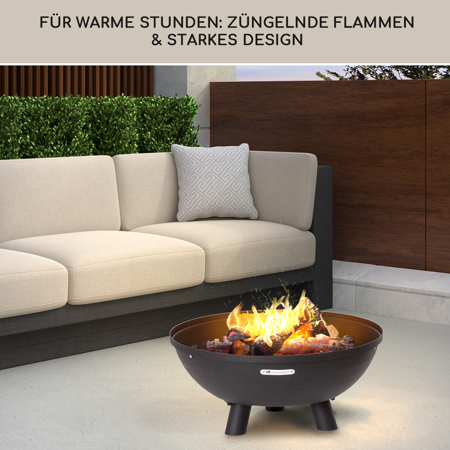 Ein schwarzer Feuerkorb mit brennendem Holz steht vor einer Couch und grünen Büschen.