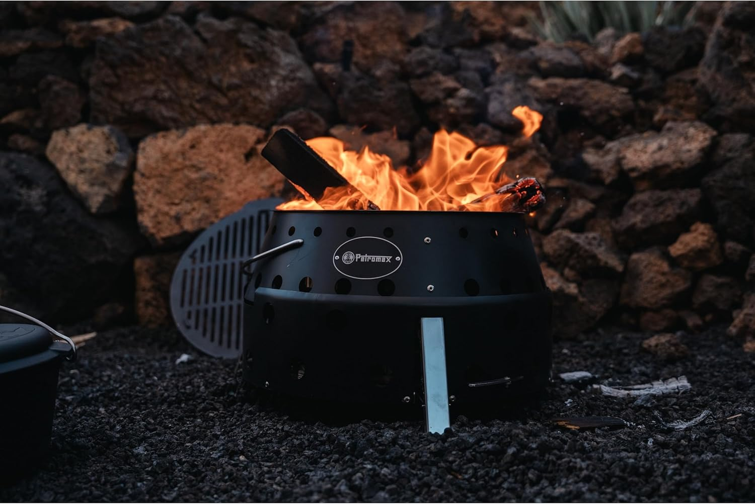 Ein schwarzer Grill mit brennendem Feuer steht auf Kies vor einer Felswand.