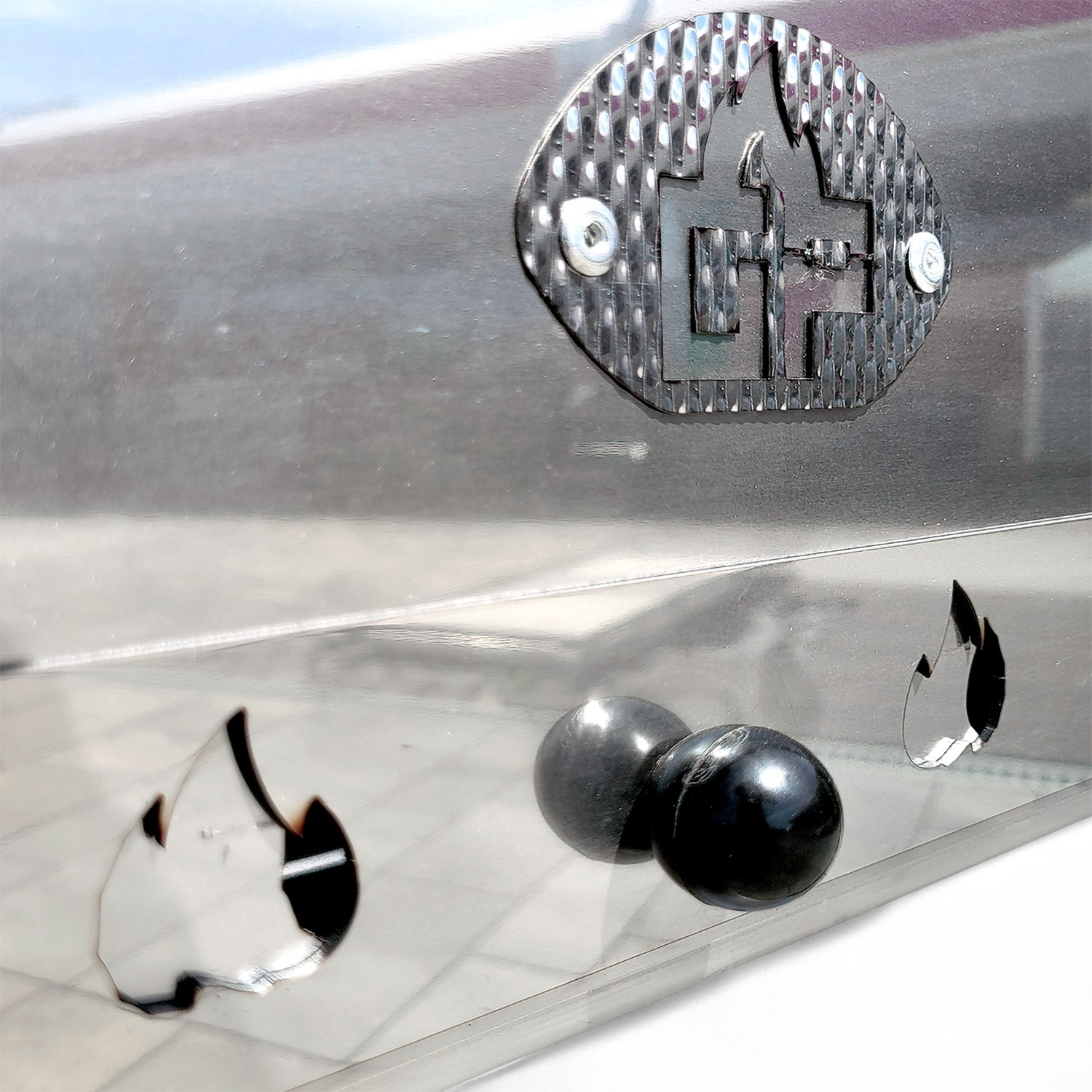 Ein metallischer Grill mit Flammenausschnitten und zwei schwarzen Knöpfen. Ein Logo ist sichtbar.