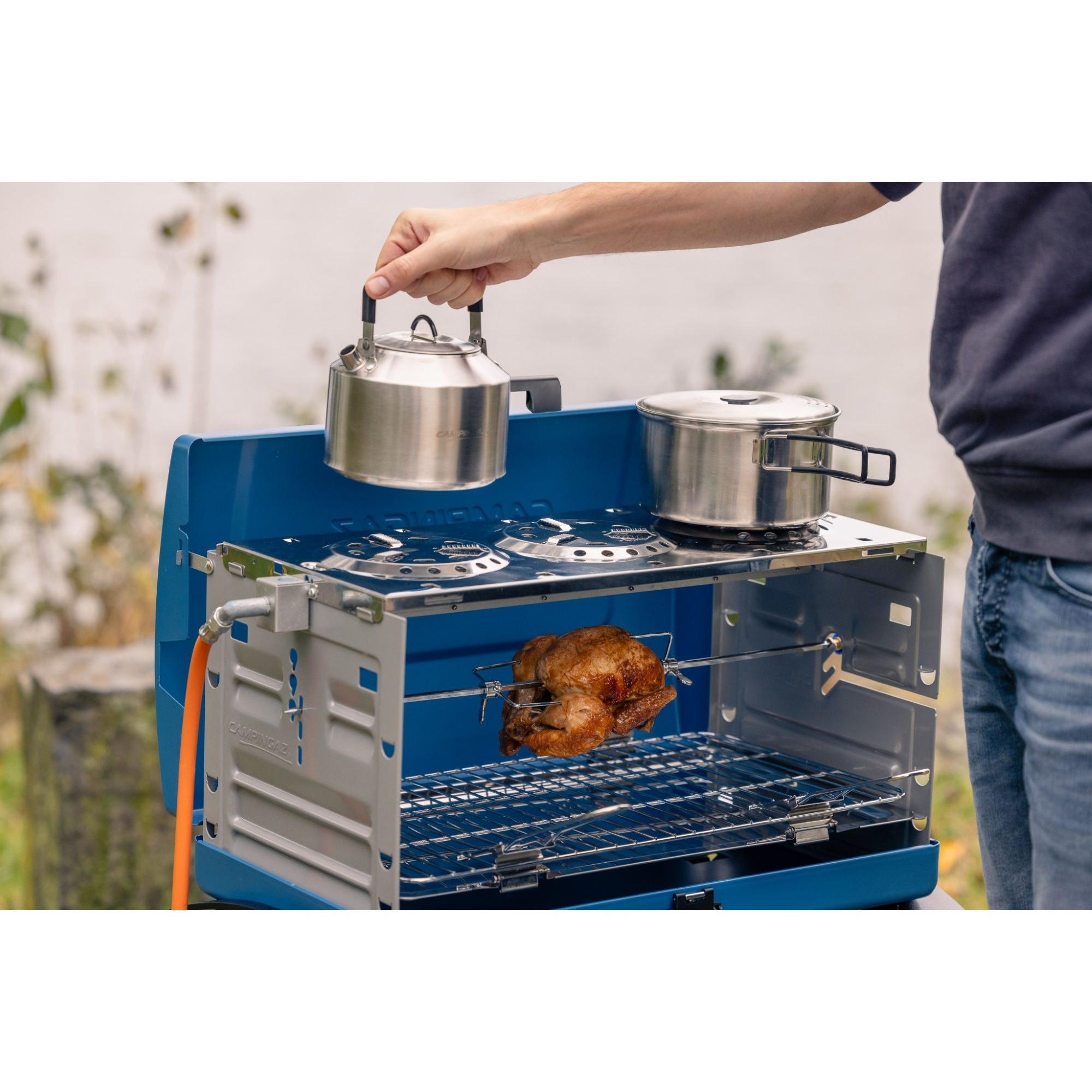 Un uomo tiene un bollitore sopra un fornello da campeggio blu con un pollo arrosto.