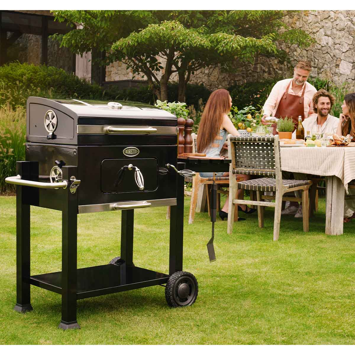 Una griglia nera in primo piano, con una famiglia che cena in giardino dietro.