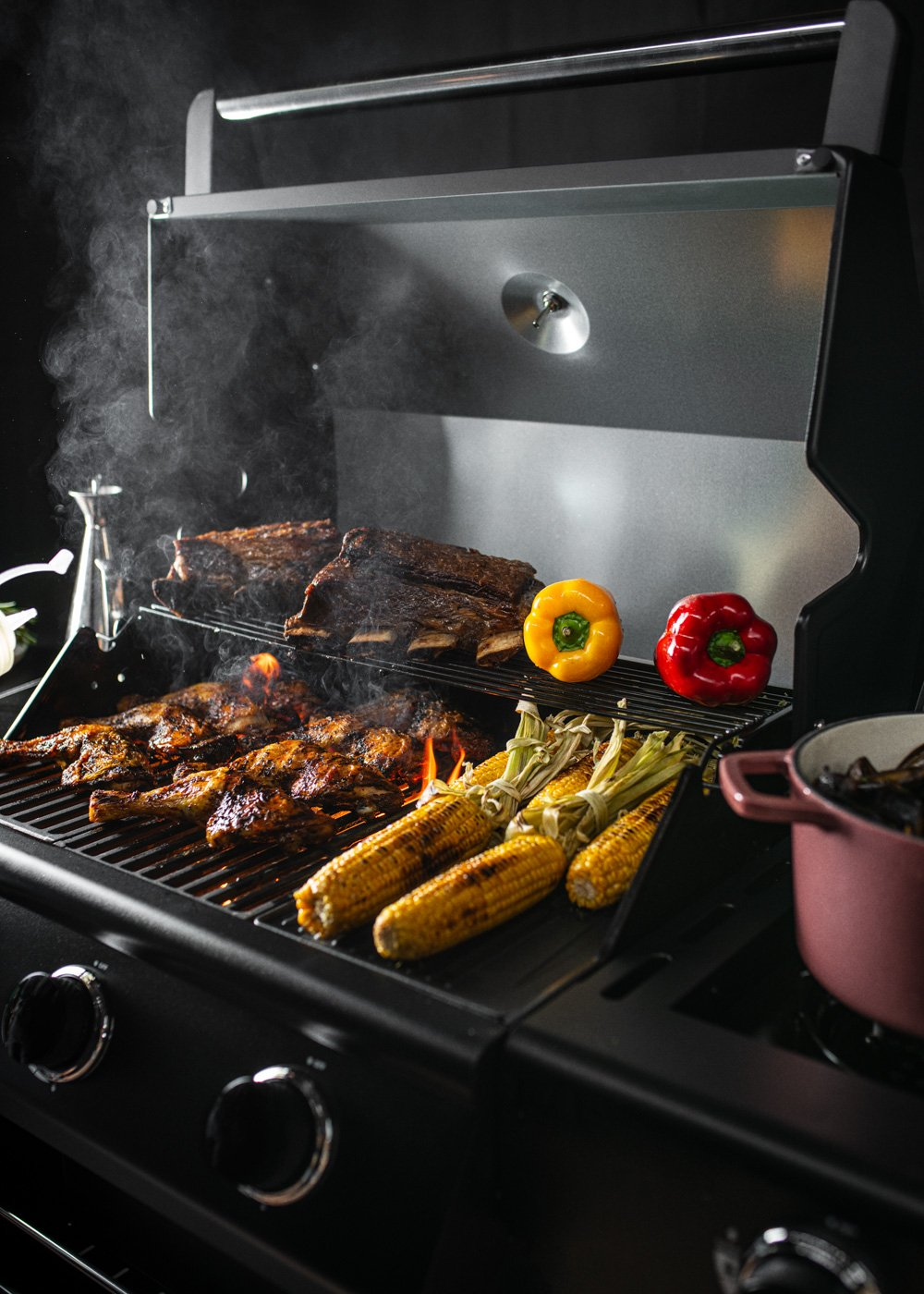 Costillas y pollo a la parrilla con maíz, pimientos y una olla al lado.