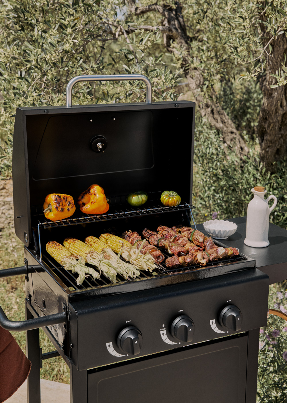 Una parrilla negra al aire libre con maíz, brochetas y pimientos asándose.