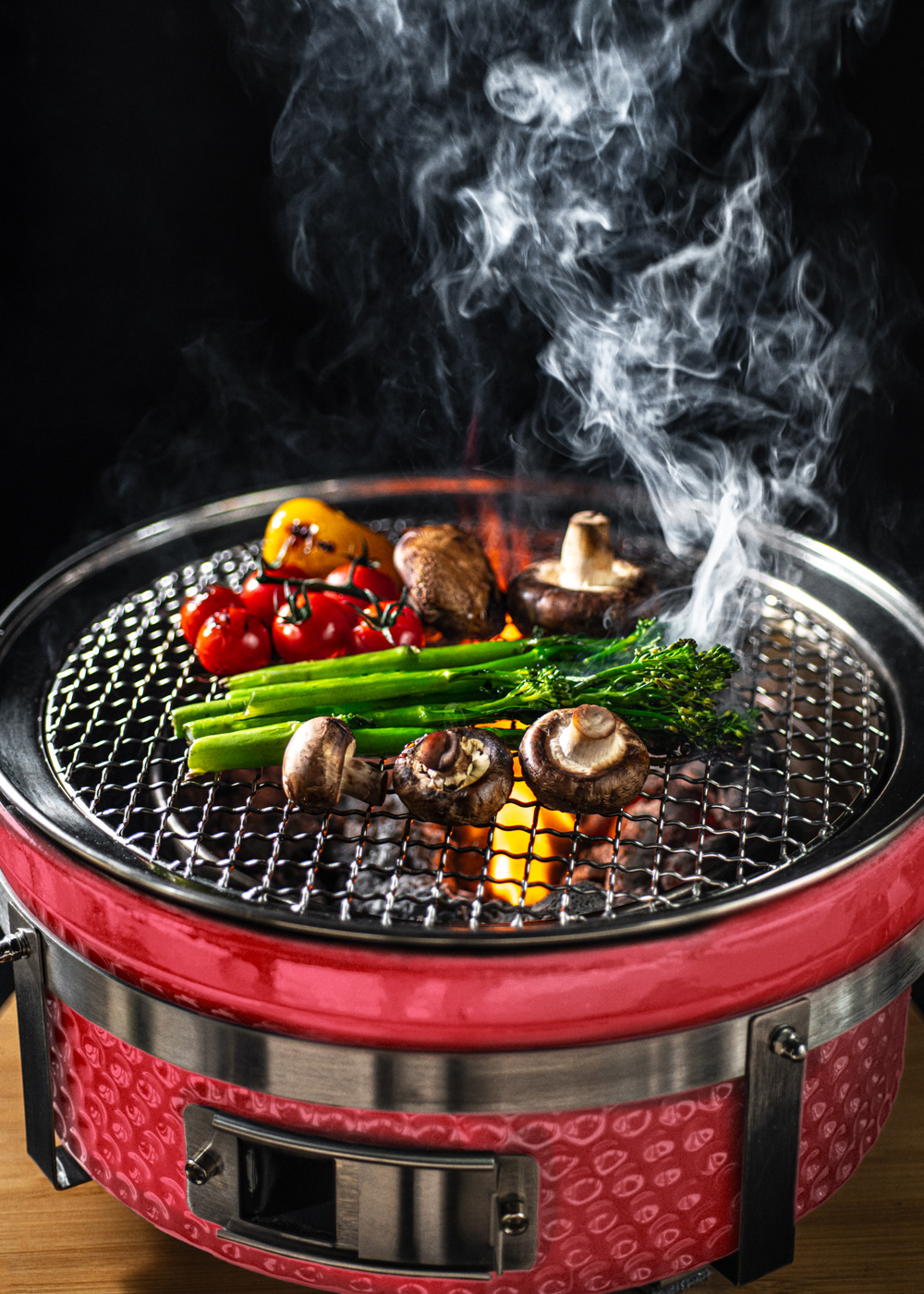 Verduras como tomates, champiñones y brócoli a la parrilla sobre una parrilla roja con llamas y humo.