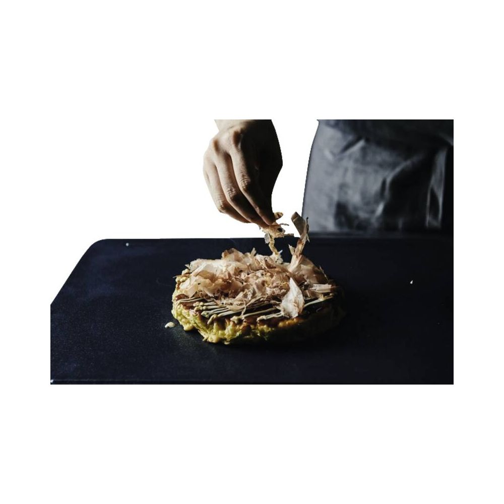 Eine Hand streut Flocken auf ein Okonomiyaki auf einer schwarzen Platte.
