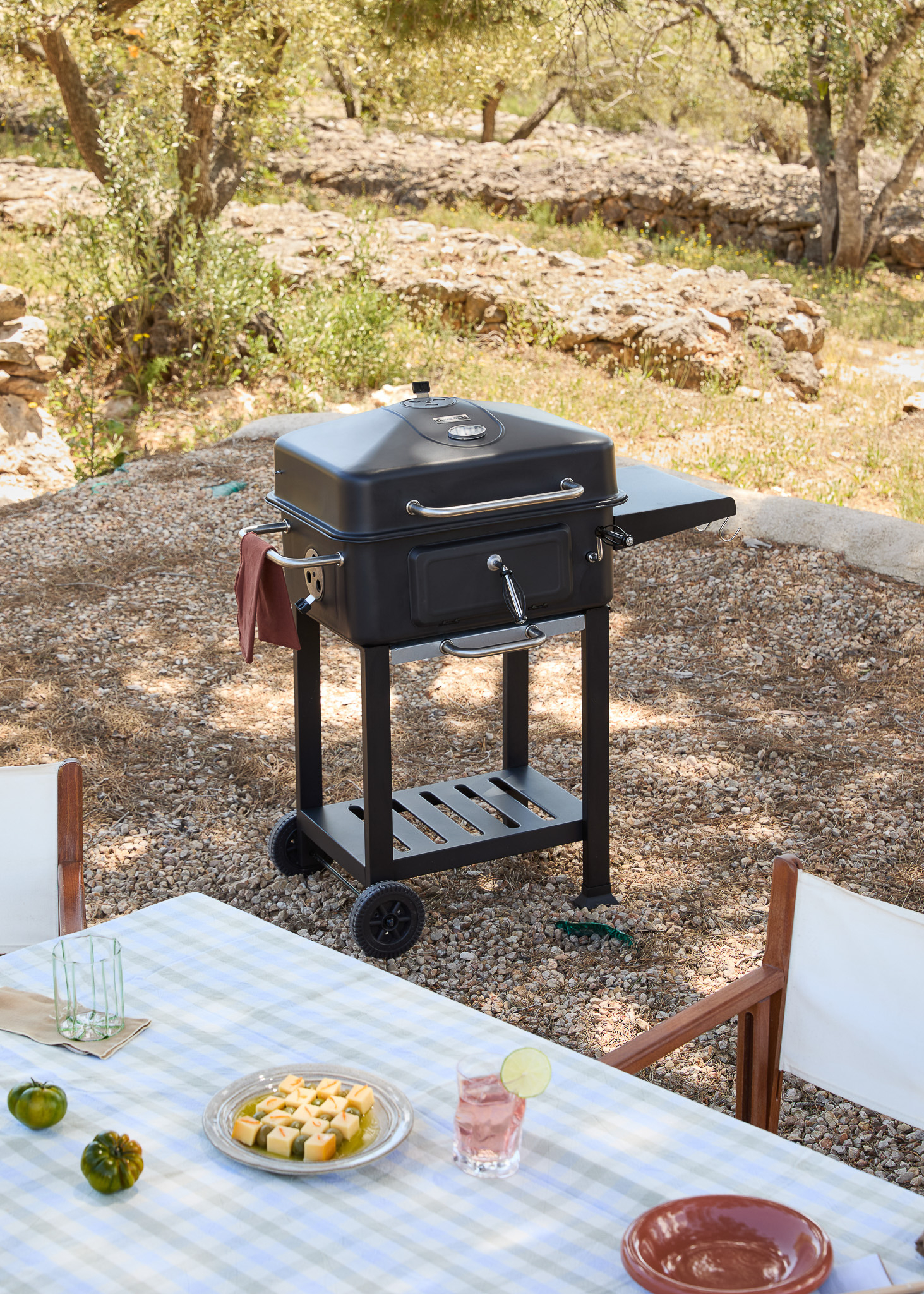 Una parrilla de carbón negra está sobre grava cerca de una mesa puesta para comer al aire libre.
