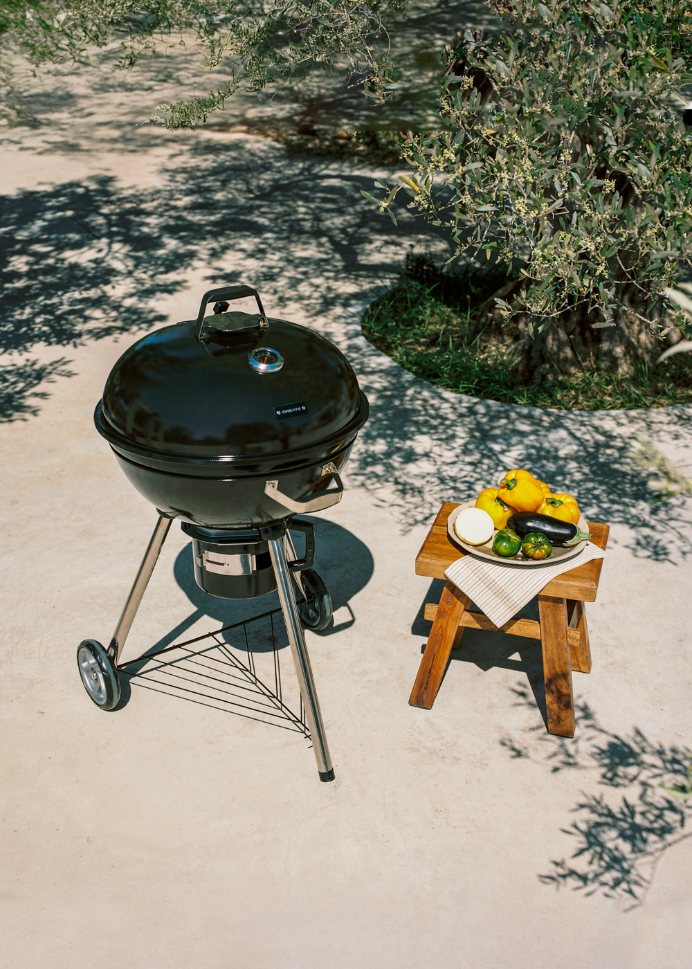 Una parrilla negra está afuera junto a un taburete de madera con verduras. Sombras de sol caen sobre el suelo.