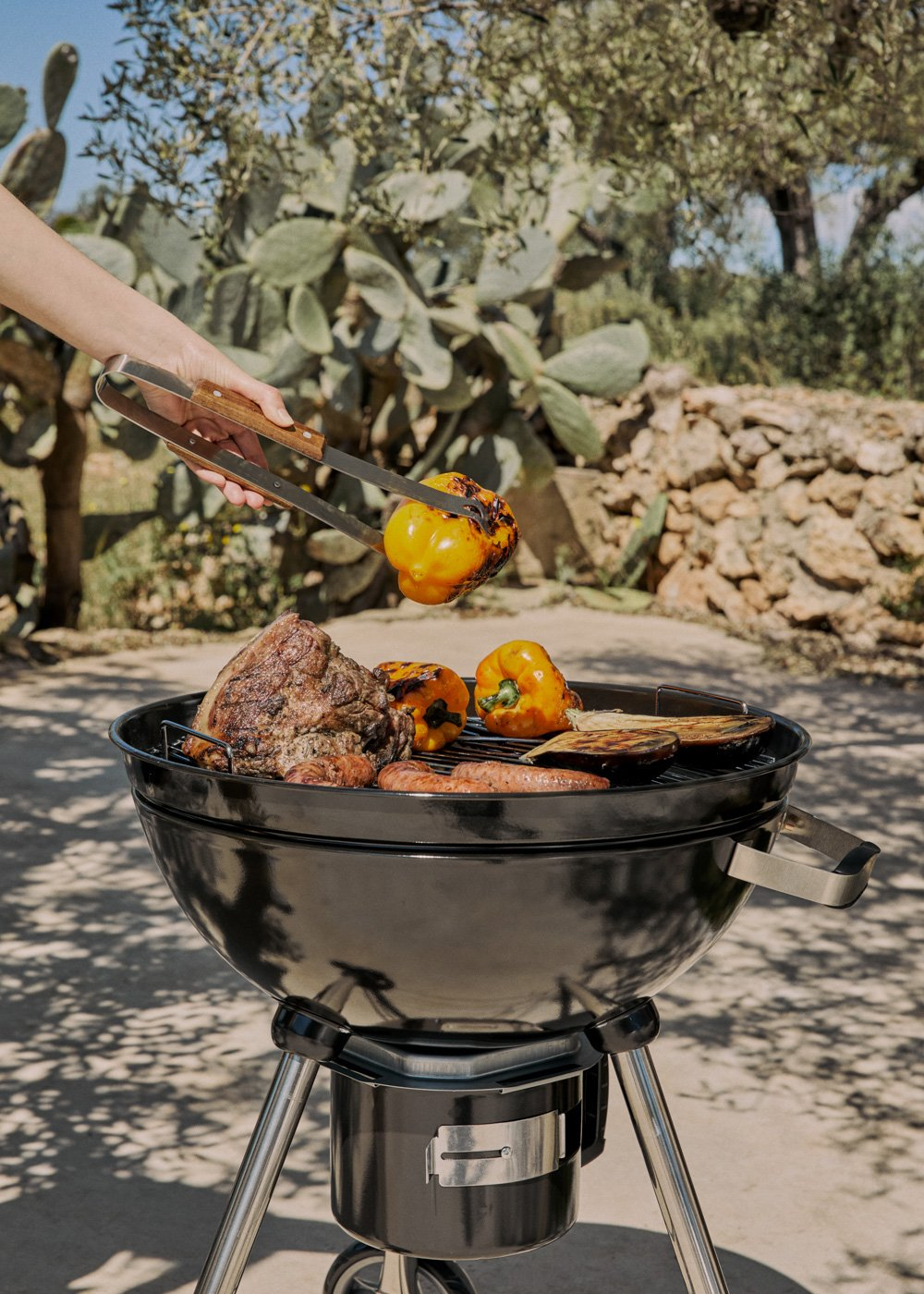 Una mano usa pinzas para colocar un pimiento amarillo en una parrilla con carne y verduras.