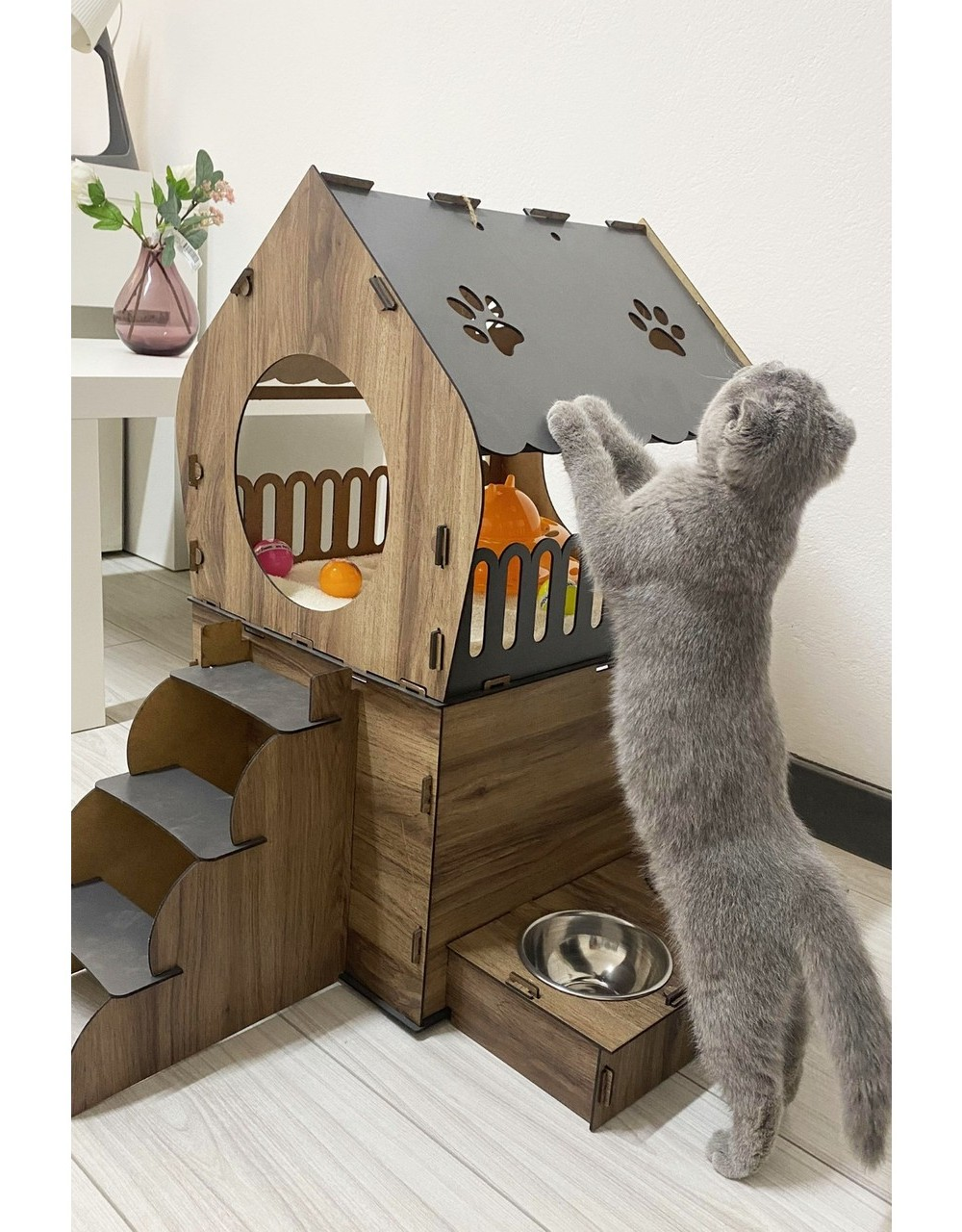 Gri bir kedi, basamaklı ahşap bir kedi evinin yanında arka ayakları üzerinde duruyor. Altında bir kase var.