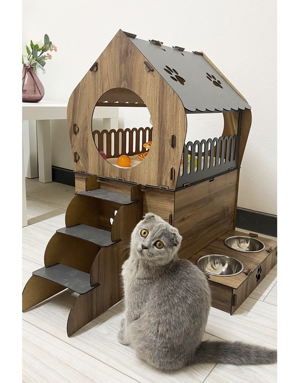 Gri bir kedi, merdivenli ve mama kaplı ahşap kedi evinin yanında oturuyor.