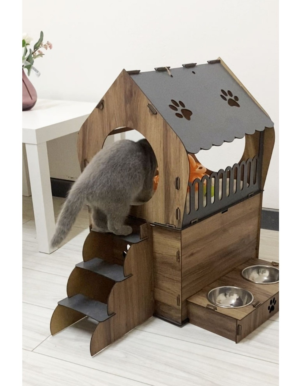 Gri bir kedi, mama kaplı ahşap kedi evine çıkan merdivenleri tırmanıyor. Çatıda pati izleri var.