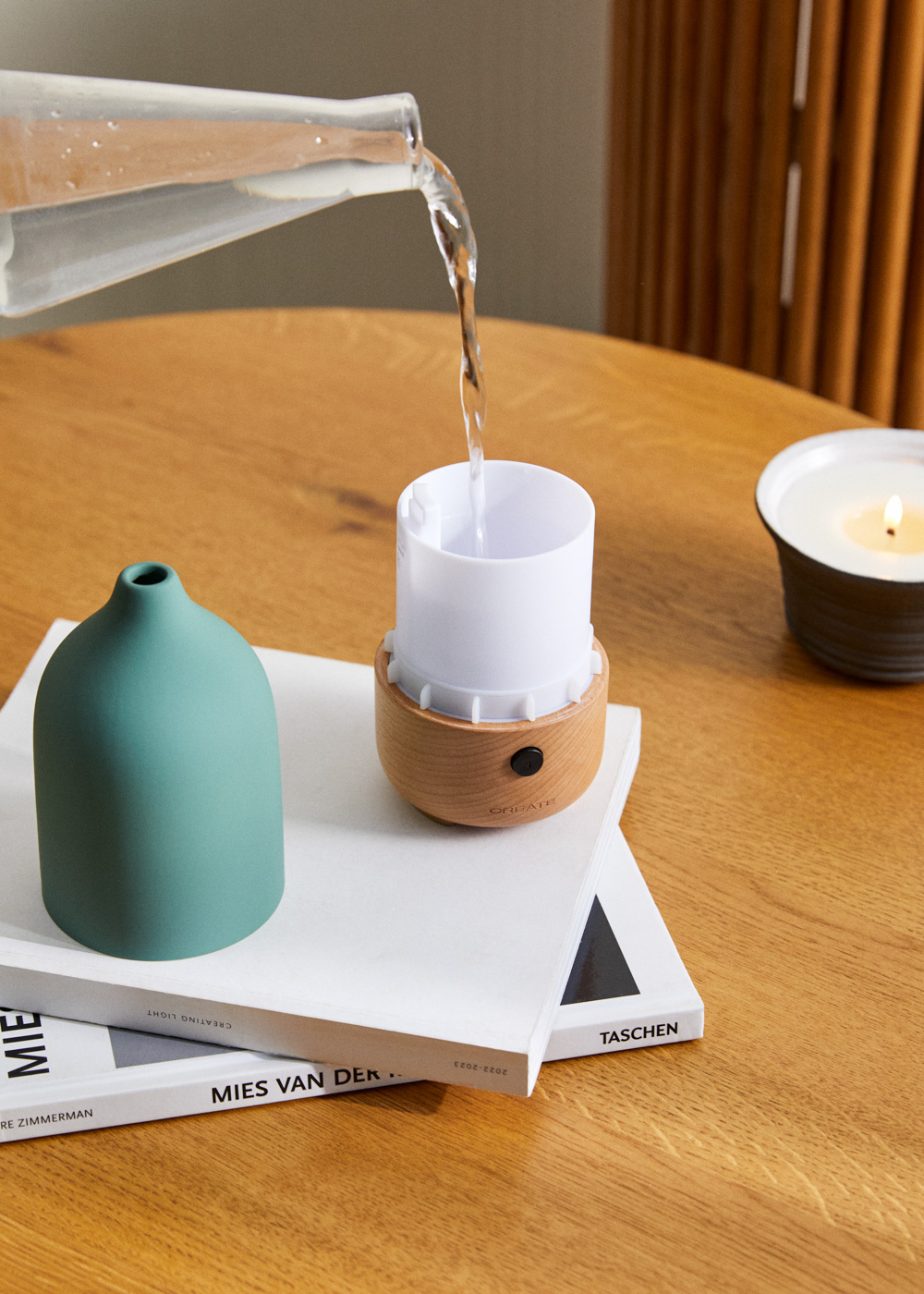 Agua se vierte de un vaso a un difusor blanco con base de madera. Cerca hay un jarrón verde y una vela.