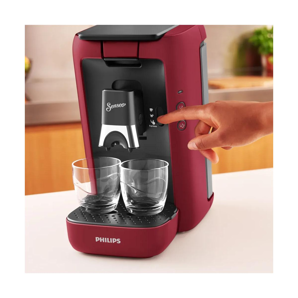 Eine rote Senseo-Kaffeemaschine mit zwei Gläsern auf einer Ablage. Eine Hand drückt einen Knopf.
