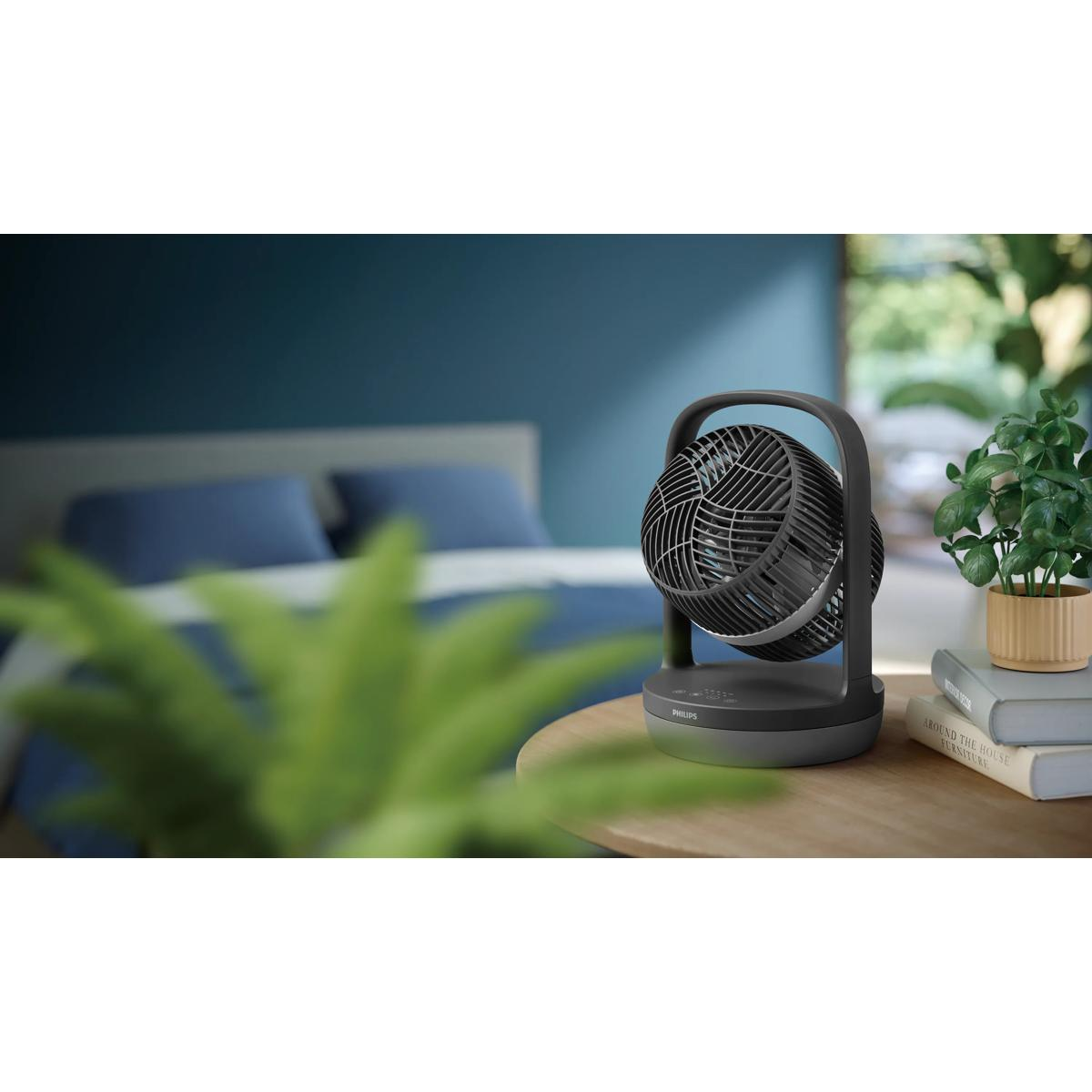 Een zwarte oscillerende ventilator staat op een houten tafel naast een potplant en boeken.