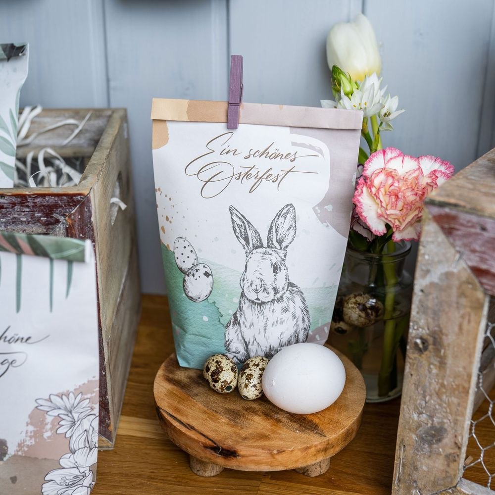 Una borsa regalo di Pasqua con stampa di coniglio e uova, accanto a una cassa di legno e fiori.