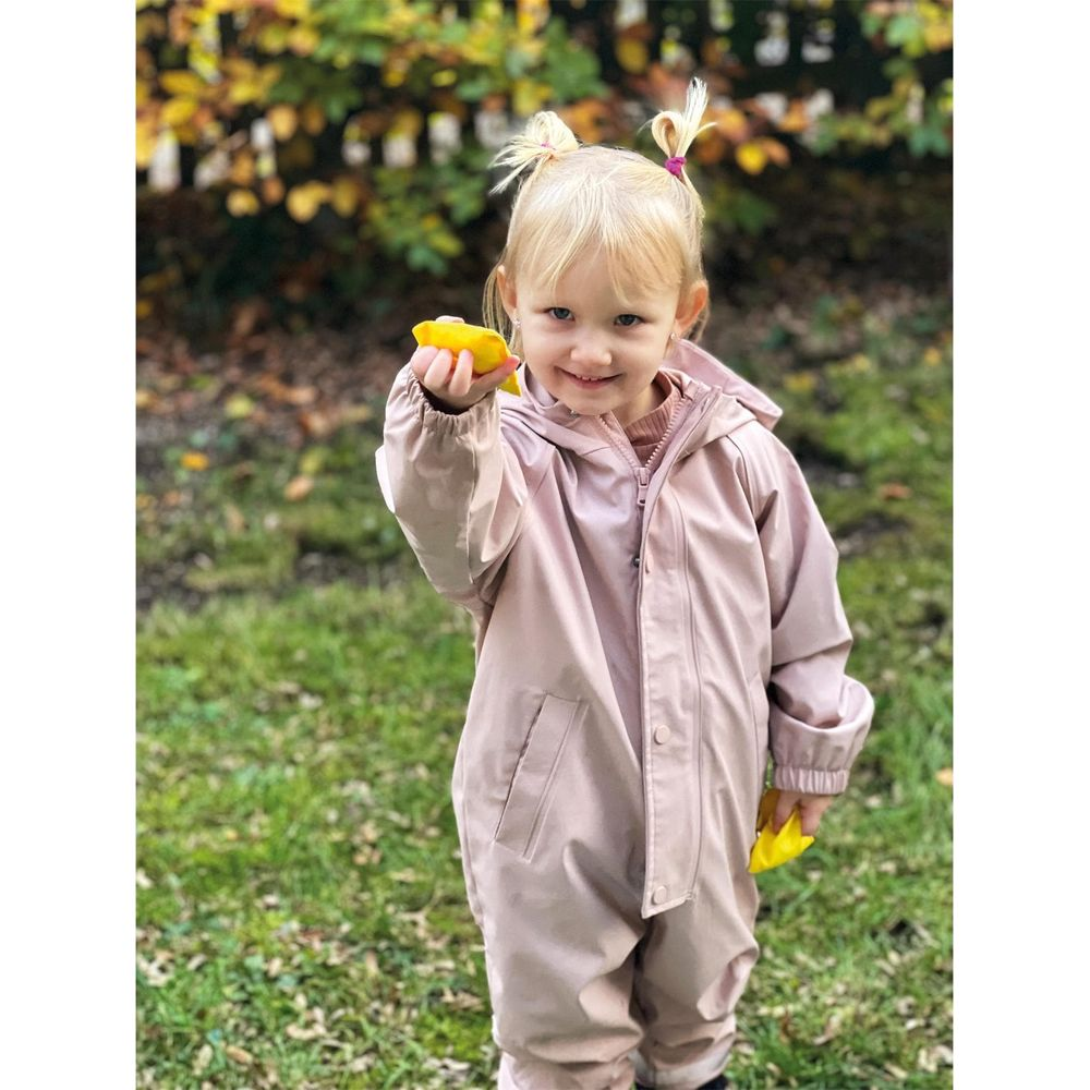 Una bambina sorridente in tuta rosa tiene oggetti gialli tra le mani all'aperto.