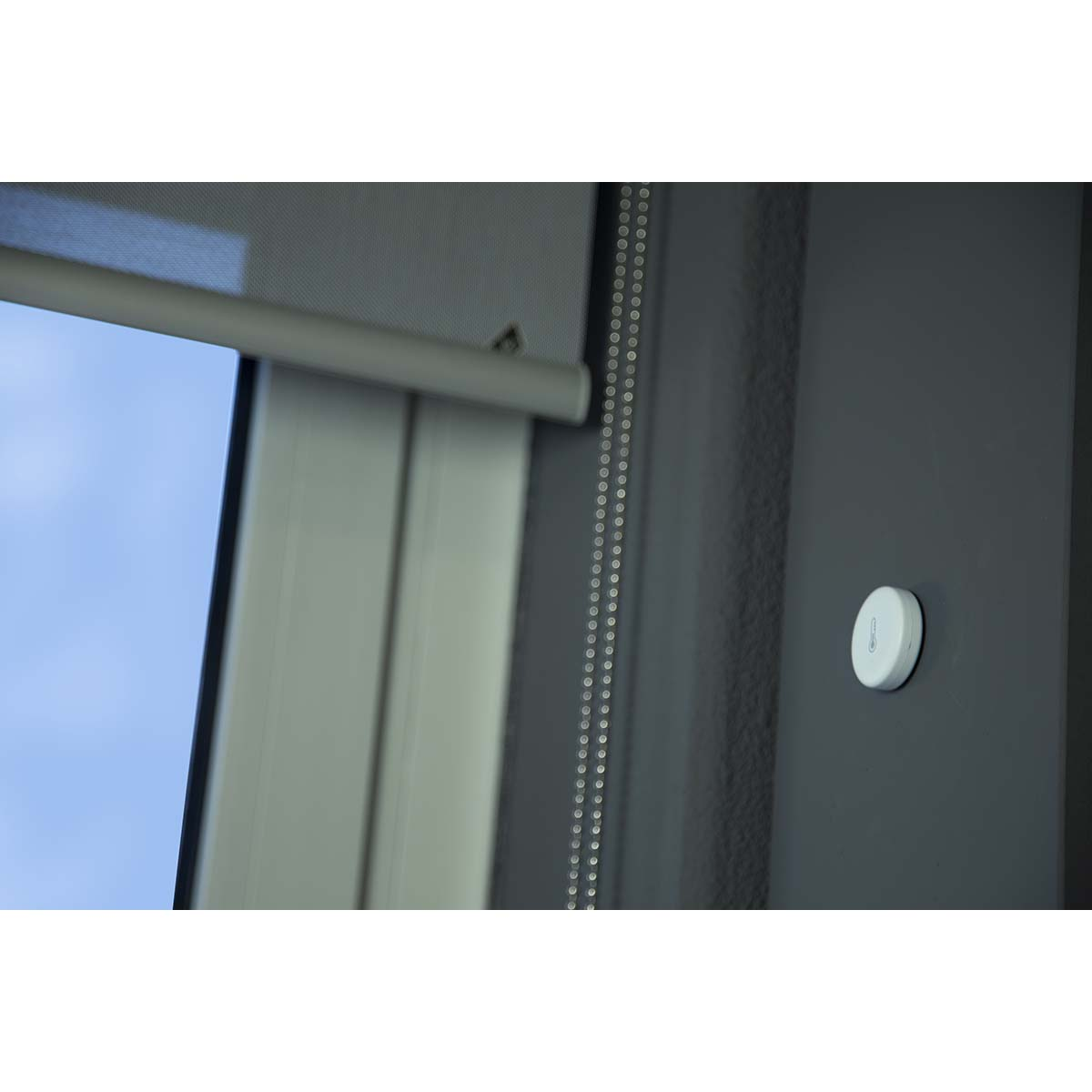 White smart home sensor on dark gray wall next to a window blind.