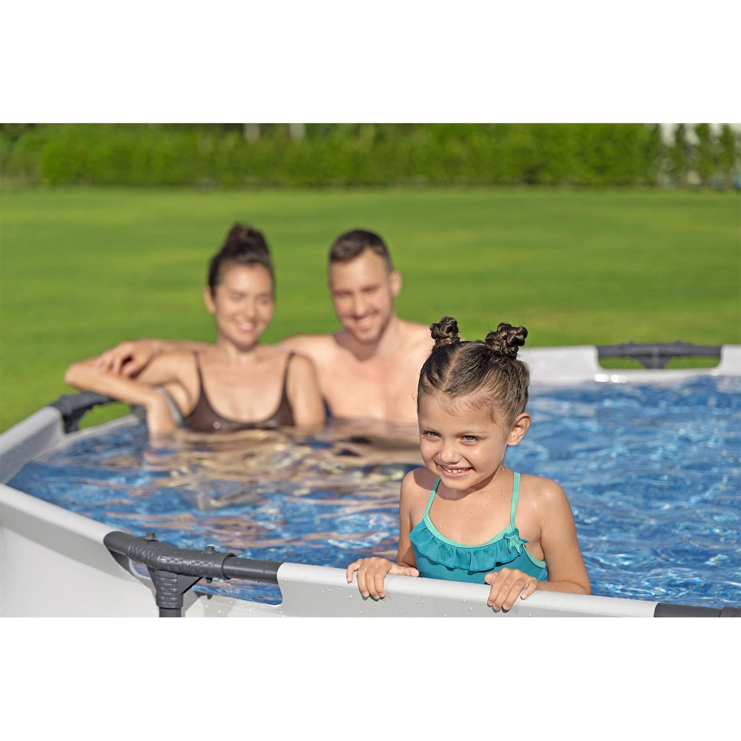 Mała dziewczynka w turkusowym stroju kąpielowym uśmiecha się w basenie. Rodzice w tle.