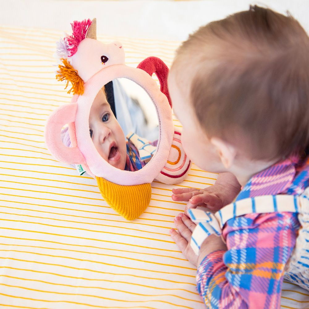 Ein Baby schaut in einen Einhorn-Spiegel auf einem gestreiften Bett.