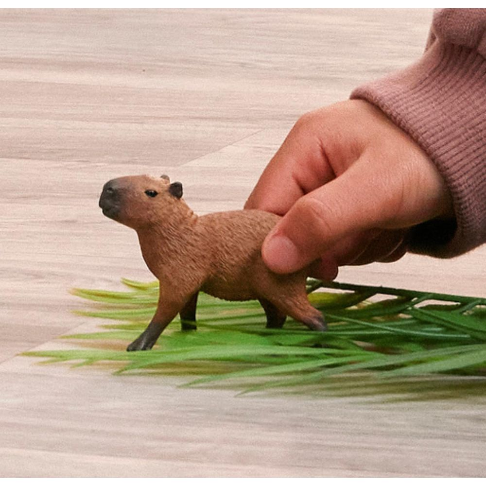 Une figurine de capybara marron sur de l'herbe verte, tenue par une main.