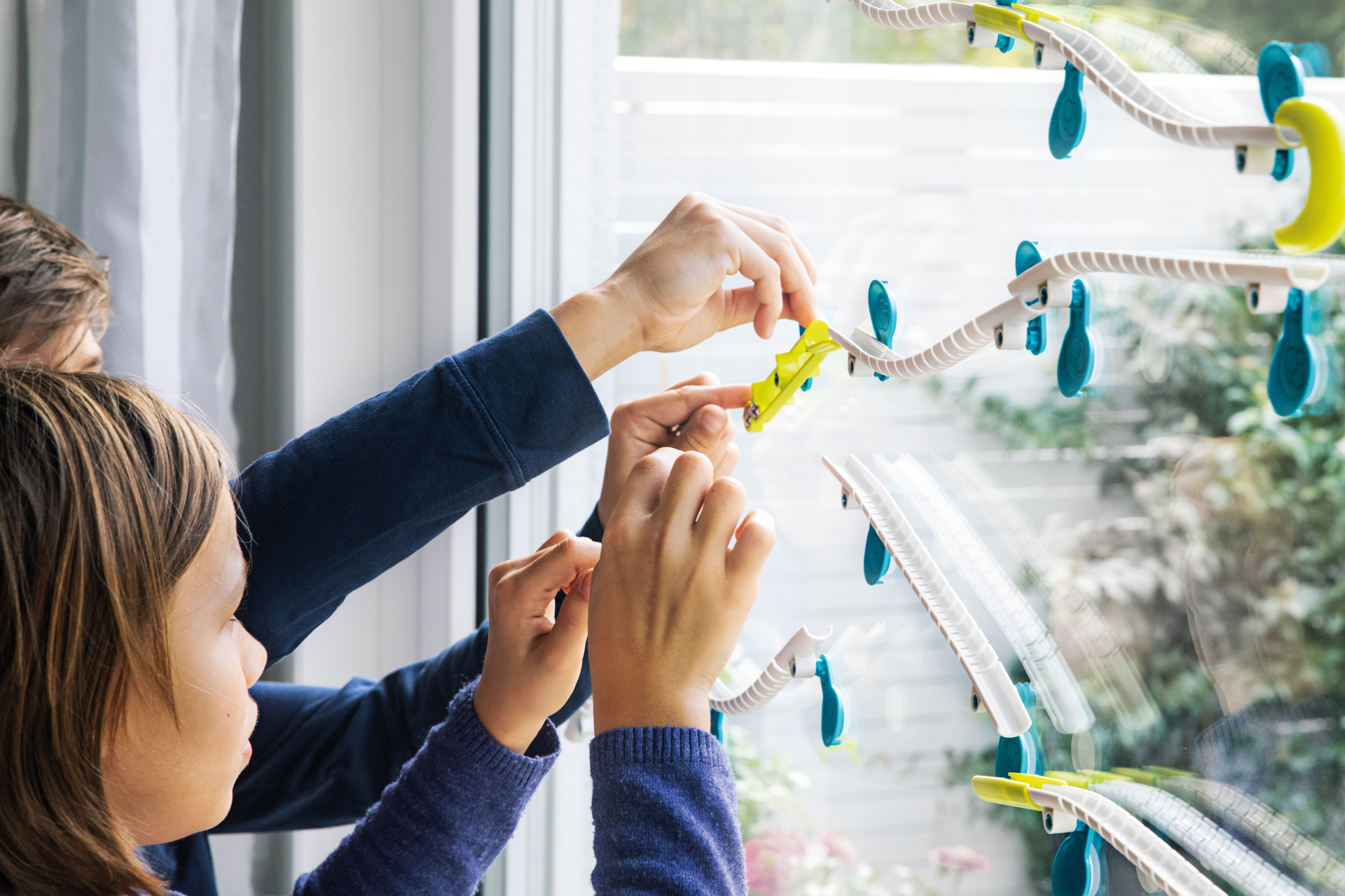 Zwei Kinder bauen eine Murmelbahn an einem Fenster. Ein gelbes Teil wird angebracht.