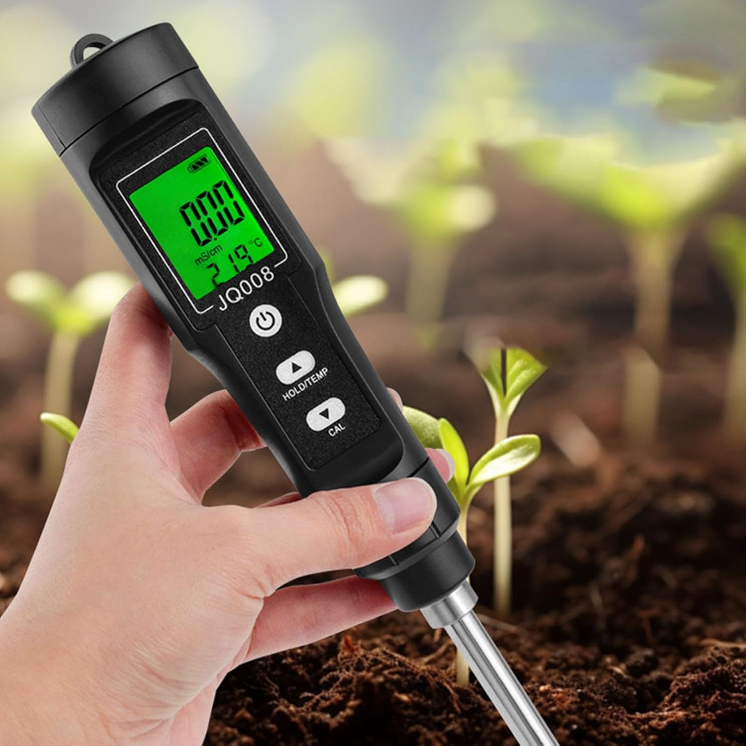 Hand hält schwarzen Bodentester mit grünem Digitaldisplay über brauner Erde und grünen Pflanzen.