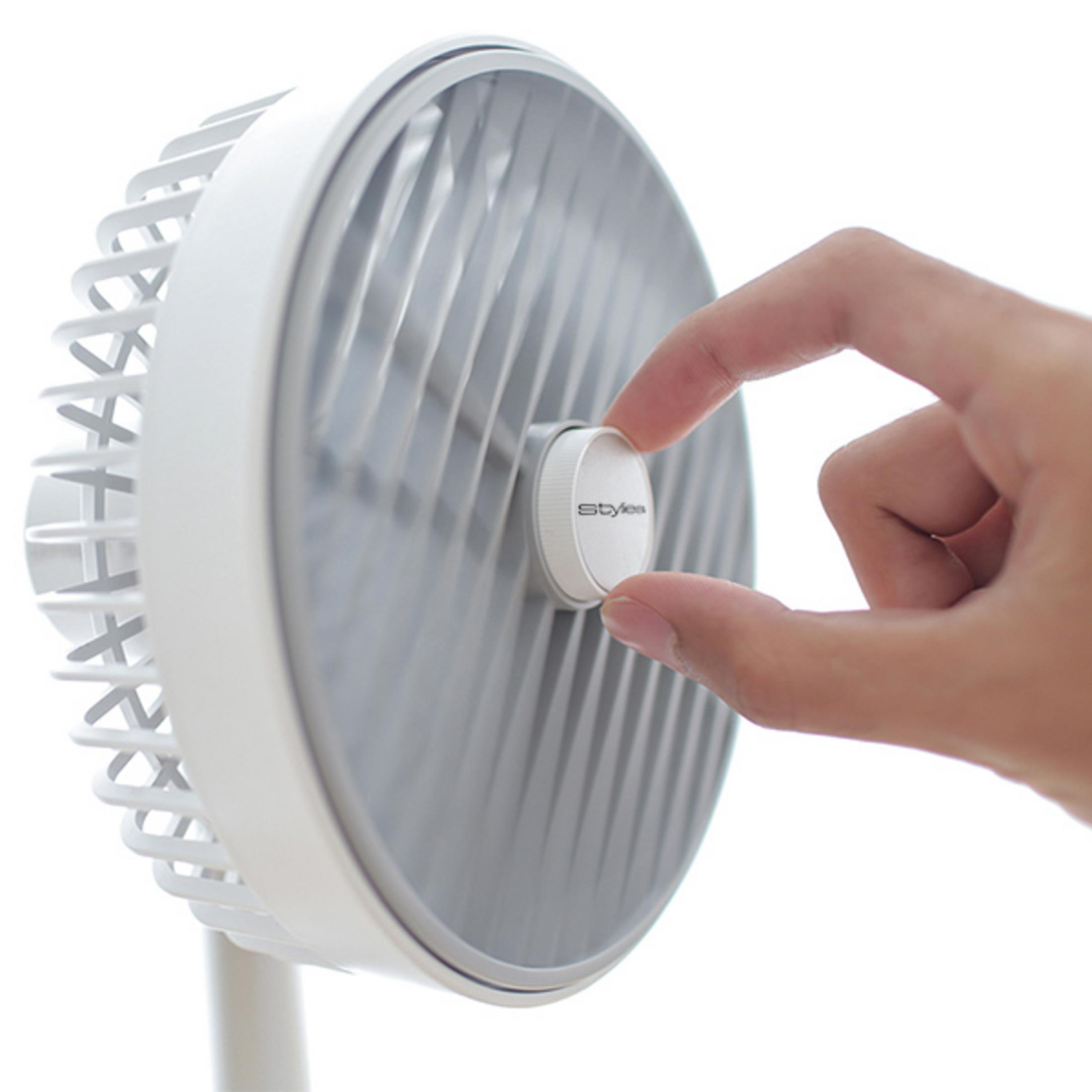 Eine Hand dreht einen weißen Knopf an einem weiß-grauen Ventilator mit geripptem Gitter.