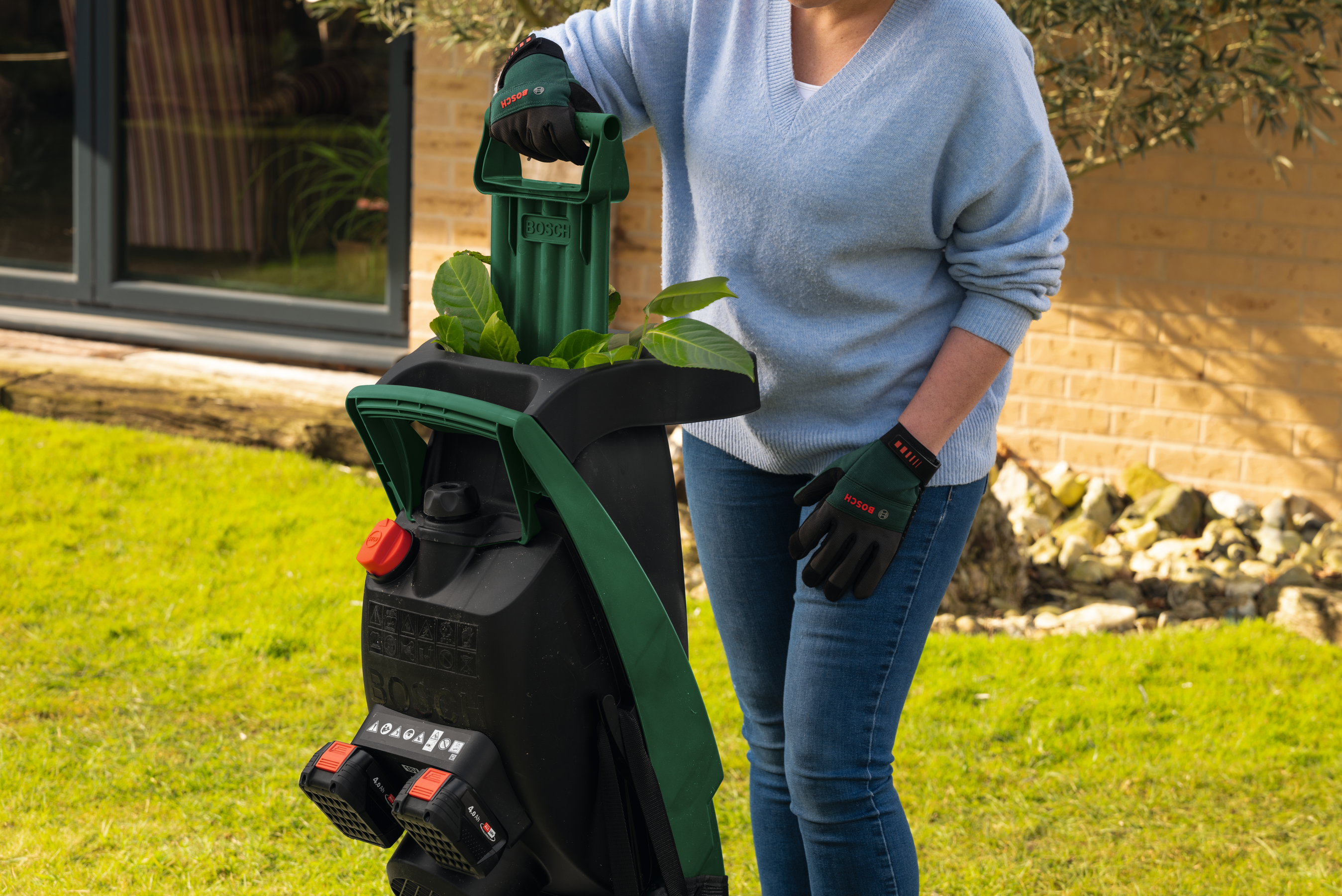 Una persona con maglione blu e jeans usa un trituratore Bosch verde in un giardino.