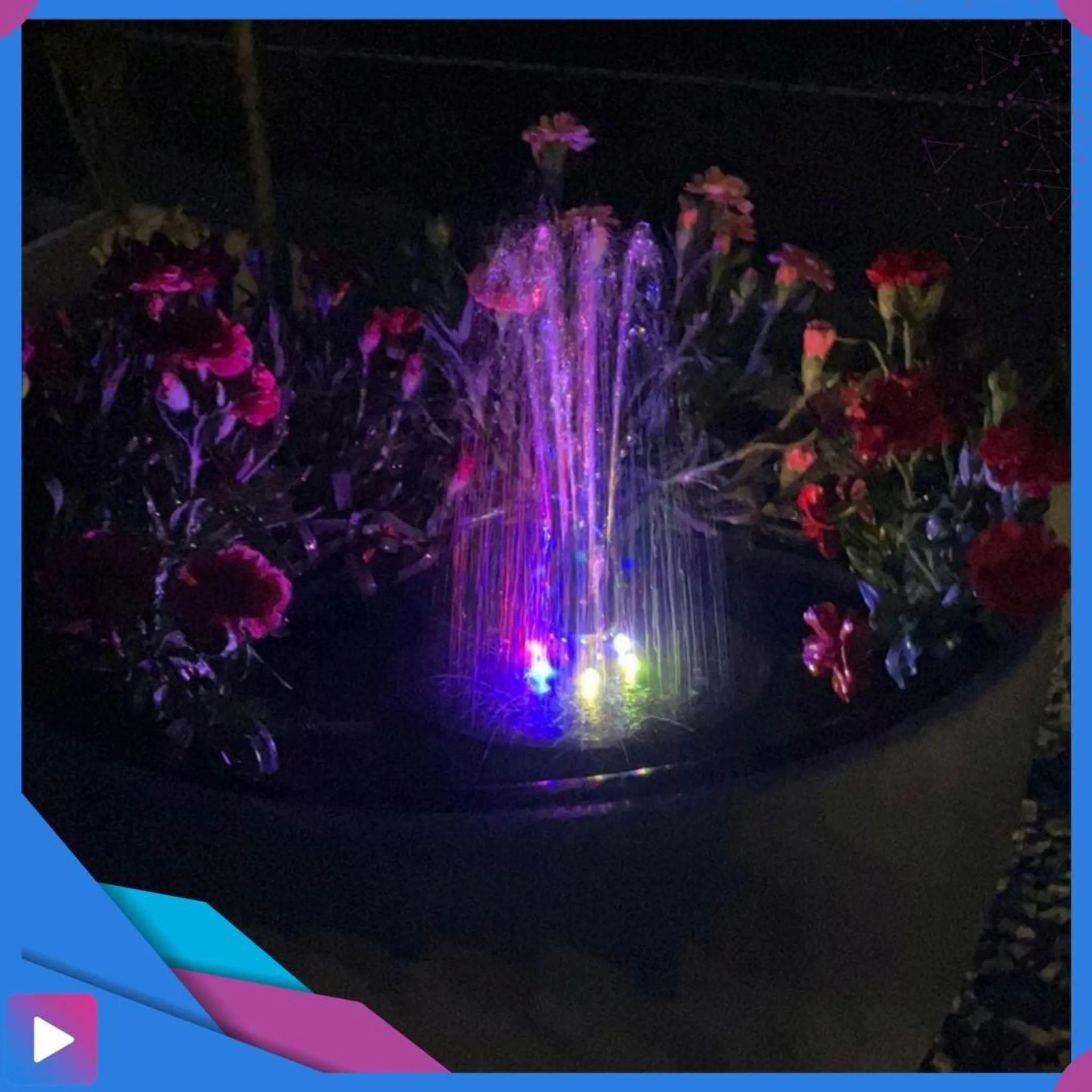 Ein kleiner beleuchteter Brunnen mit bunten Lichtern in einem Blumenbeet.