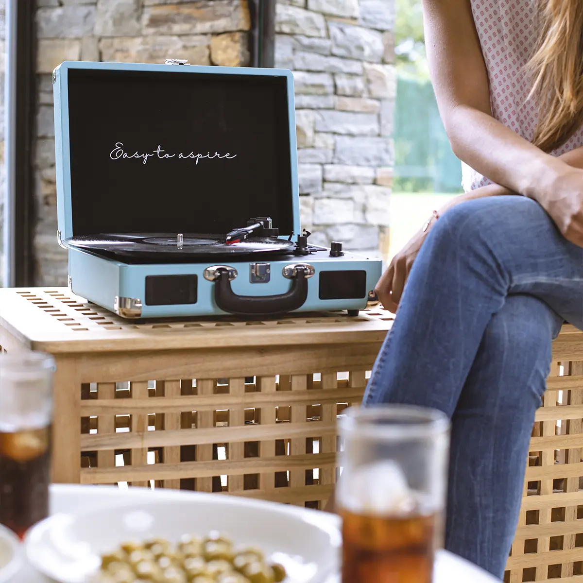 Jasnoniebieski gramofon z otwartą pokrywą stoi na drewnianym stole. Obok siedzi osoba.