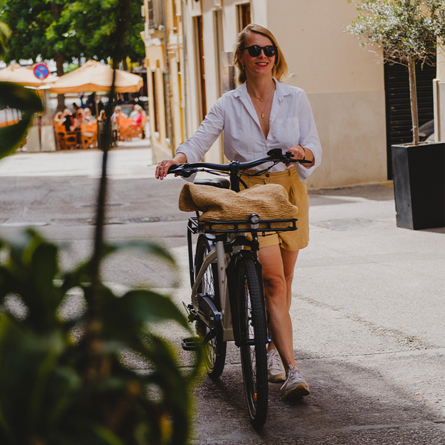 Eine Frau mit Sonnenbrille und weißem Hemd schiebt ein Fahrrad mit Strohkorb.
