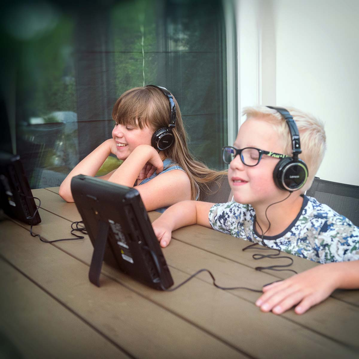 Zwei Kinder sitzen draußen an einem Tisch und hören über Kopfhörer Musik.
