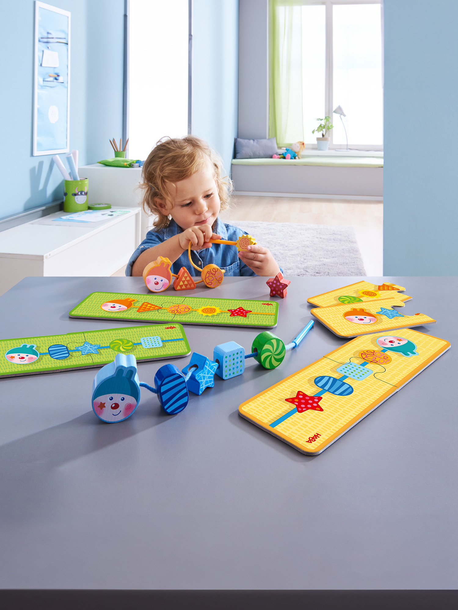 Ein kleines Kind fädelt bunte Holzperlen auf eine Schnur, Spielzeug liegt auf dem Tisch.