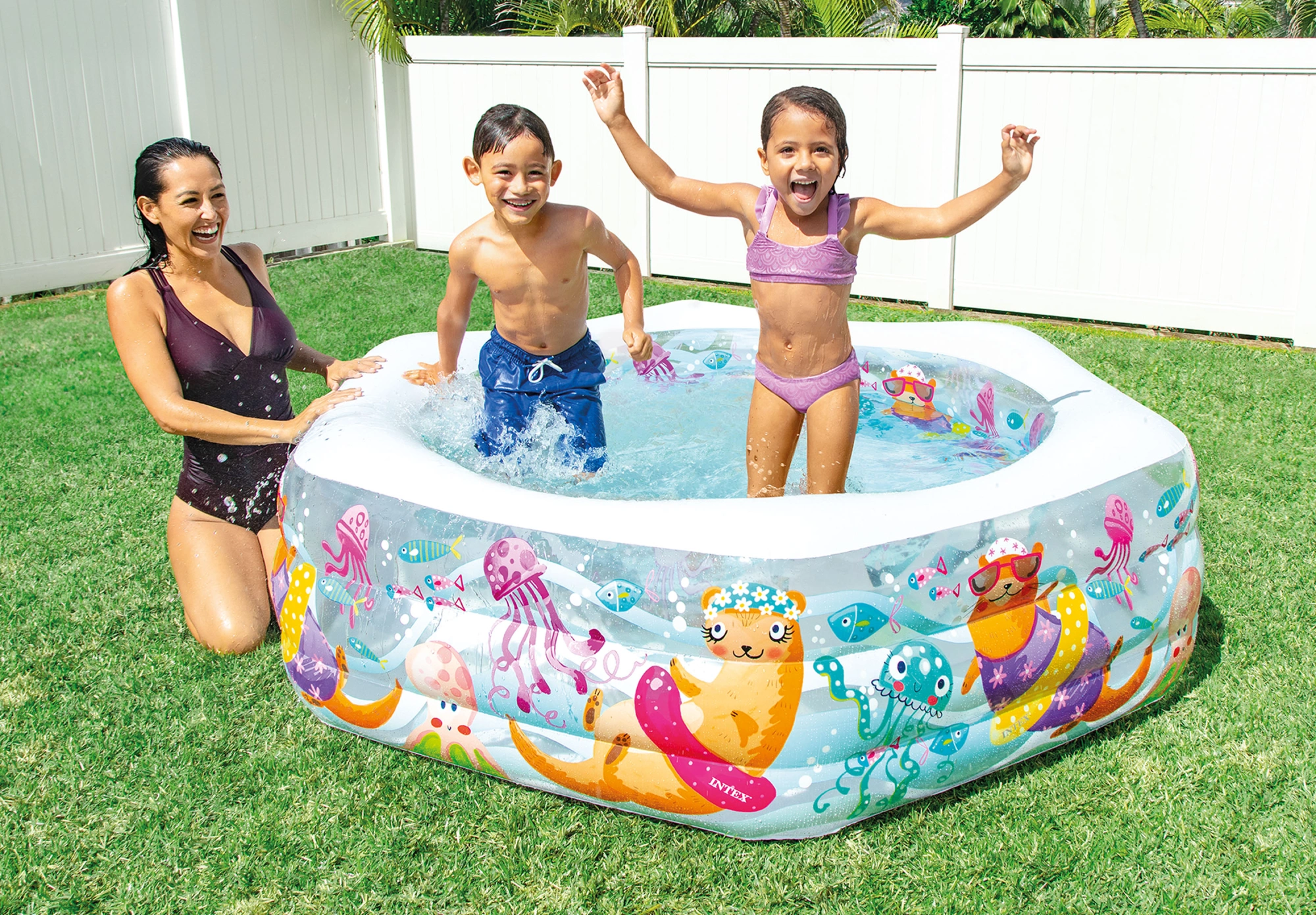 Una donna e due bambini sguazzano gioiosamente in una piscina gonfiabile decorata con colorate creature marine su un prato soleggiato.