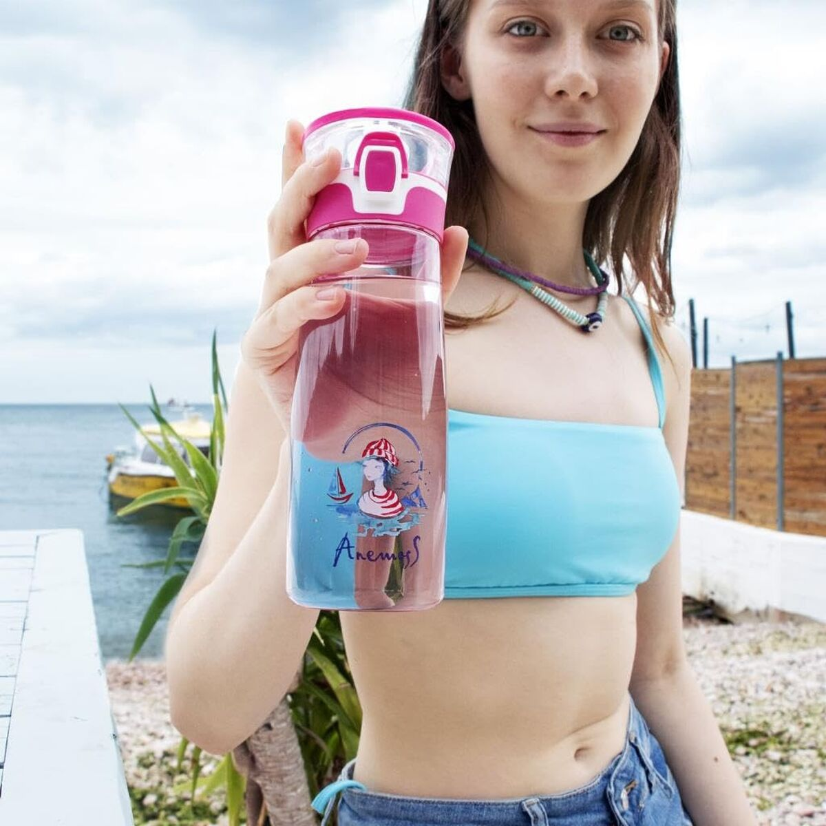Kadın plajda 'Anemoss' yazılı, deniz grafikli pembe su şişesi tutuyor.