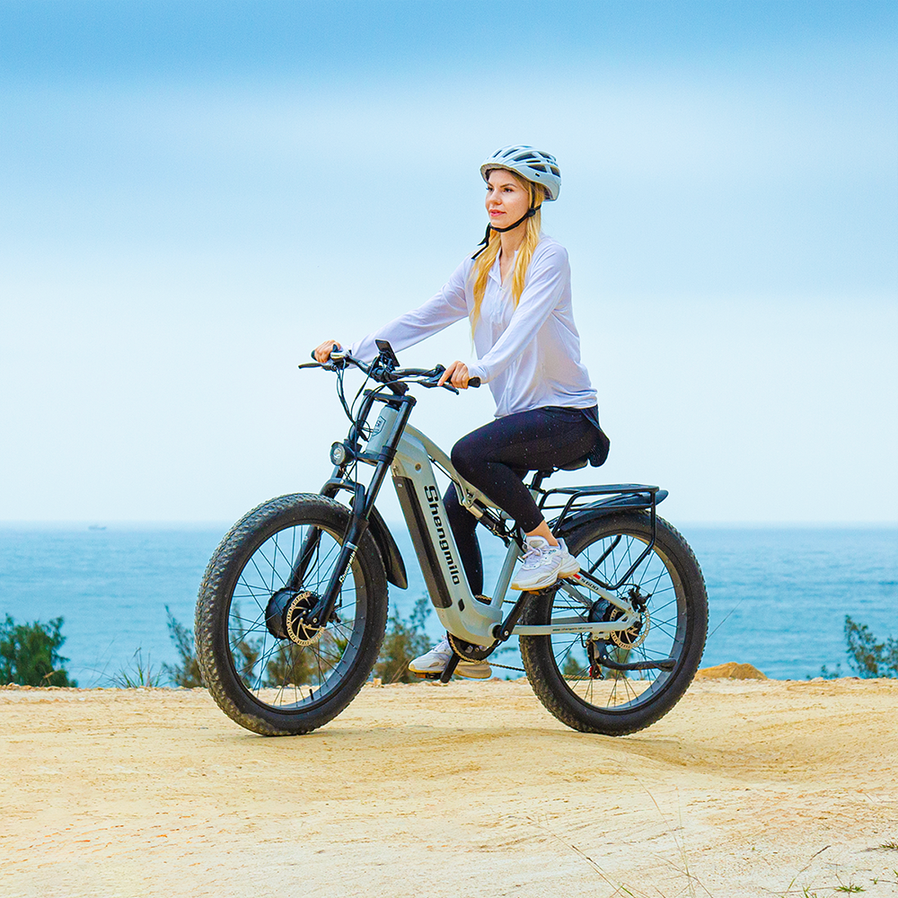 Frau mit Helm fährt ein graues E-Bike auf sandigem Weg am Meer.